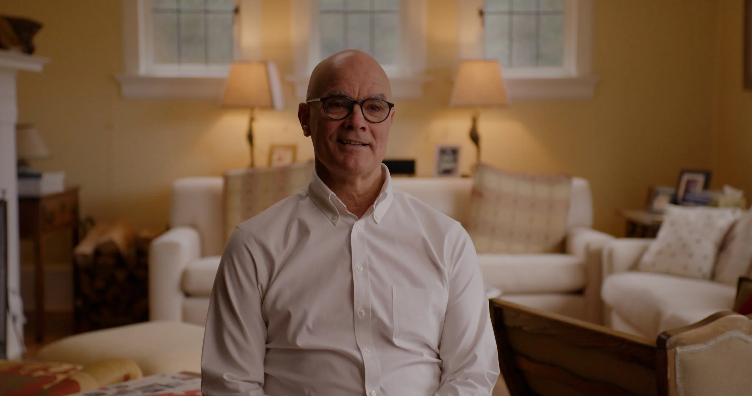 An elderly man with glasses and a white dress shirt sits in a cozy living room with beige walls, sofas, and lamps.