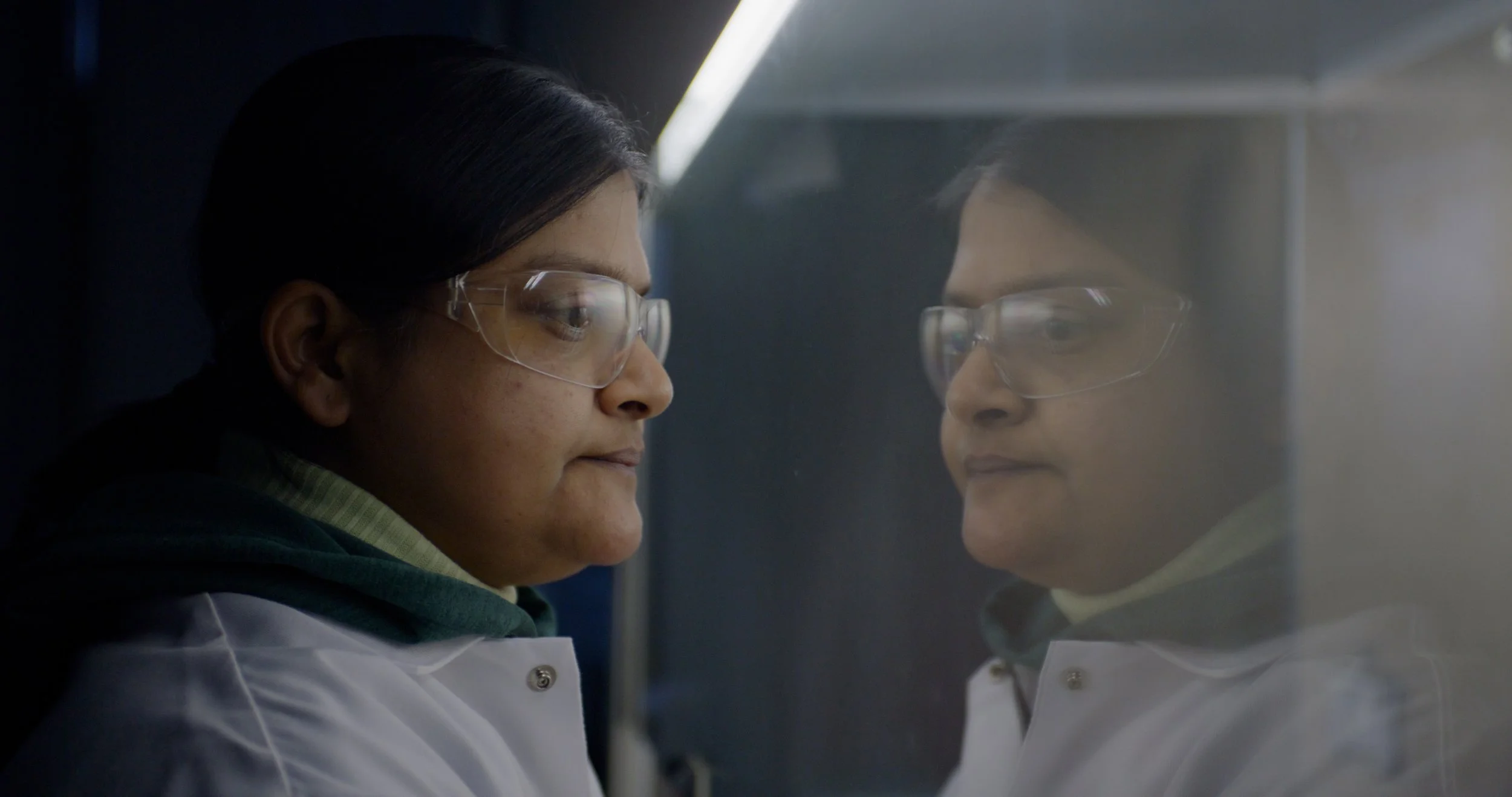 A scientist wearing safety glasses and a lab coat looks at her reflection on a glass surface in a laboratory setting.