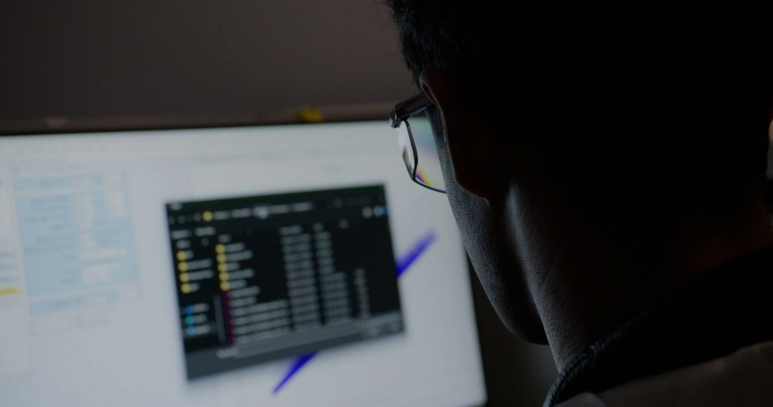 Silhouette of a person wearing glasses working at a computer with a blurred screen displaying data or code.