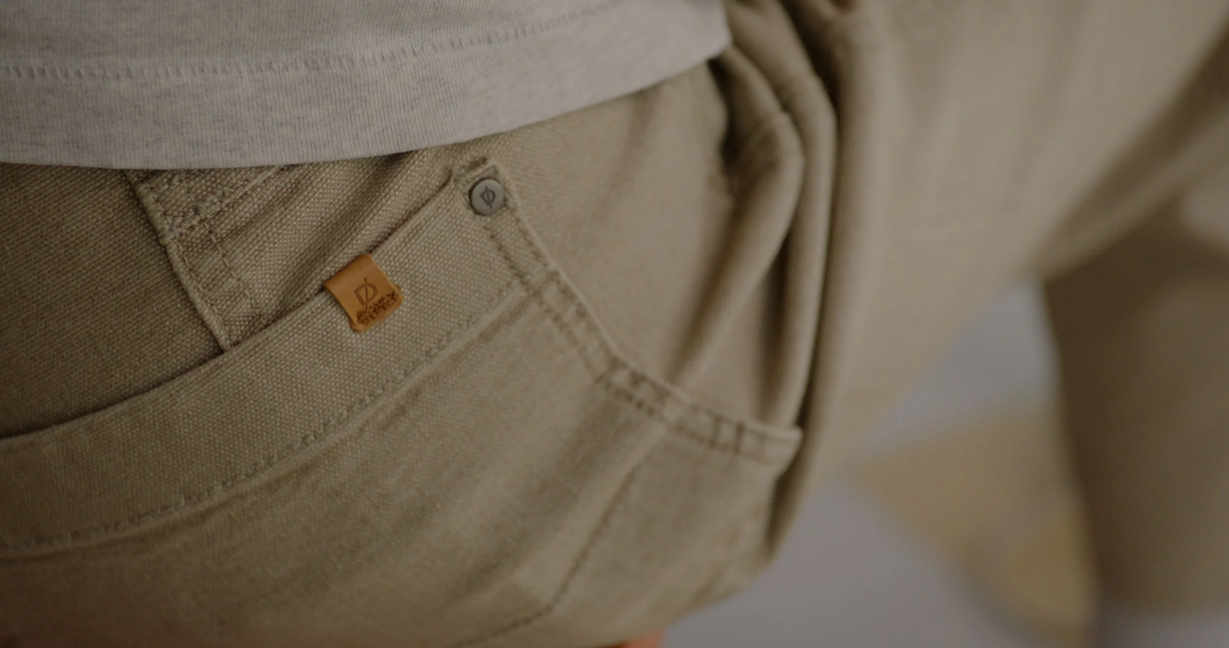 Close-up of beige pants with an orange Brand logo tag and a metal button, a portion of a white shirt is visible at the top.