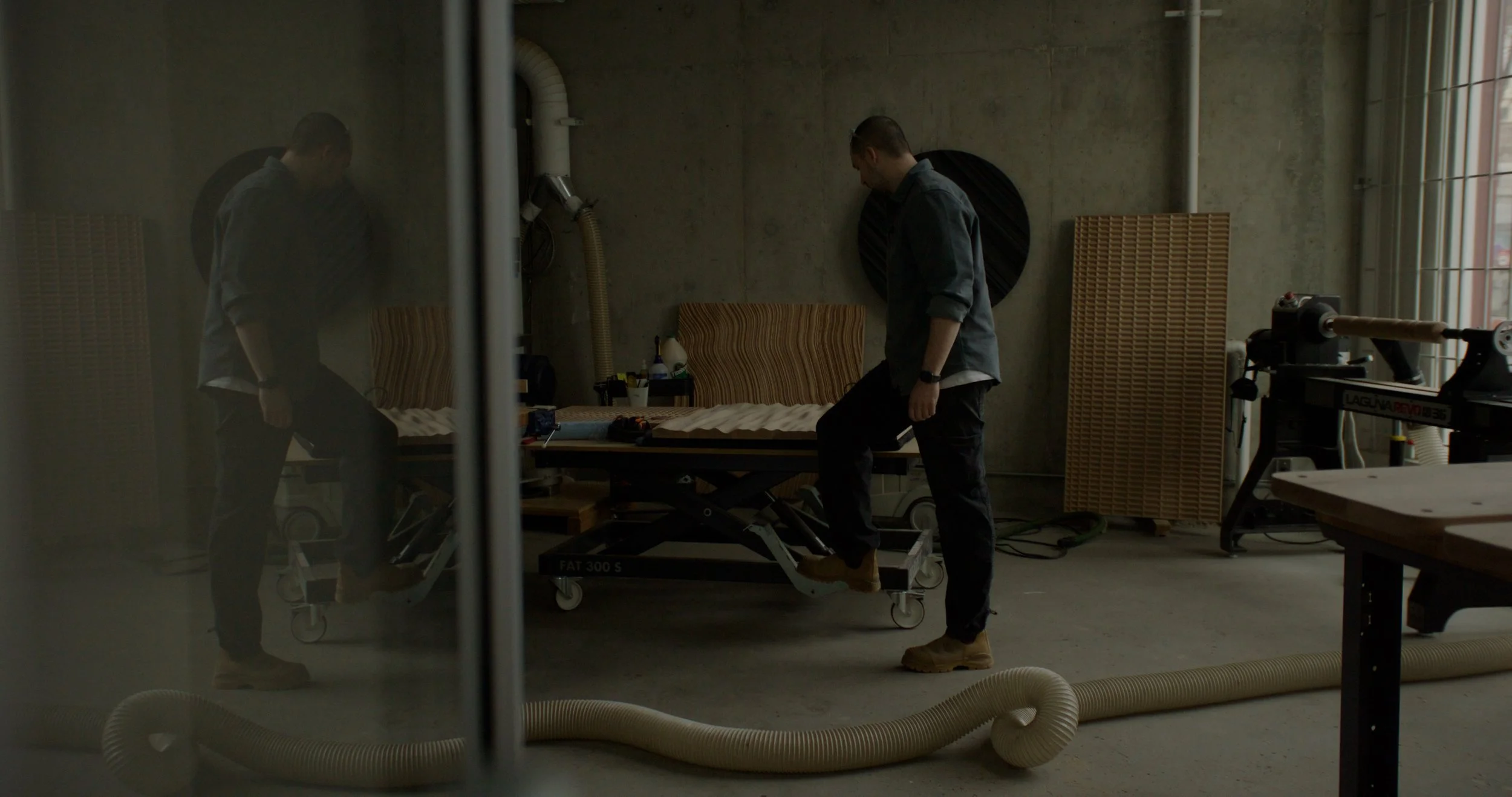 Two men in a dimly lit industrial-style room standing next to a medical stretcher, appearing to be adjusting it or inspecting it. The room has concrete walls, various equipment, and a large hose on the floor.