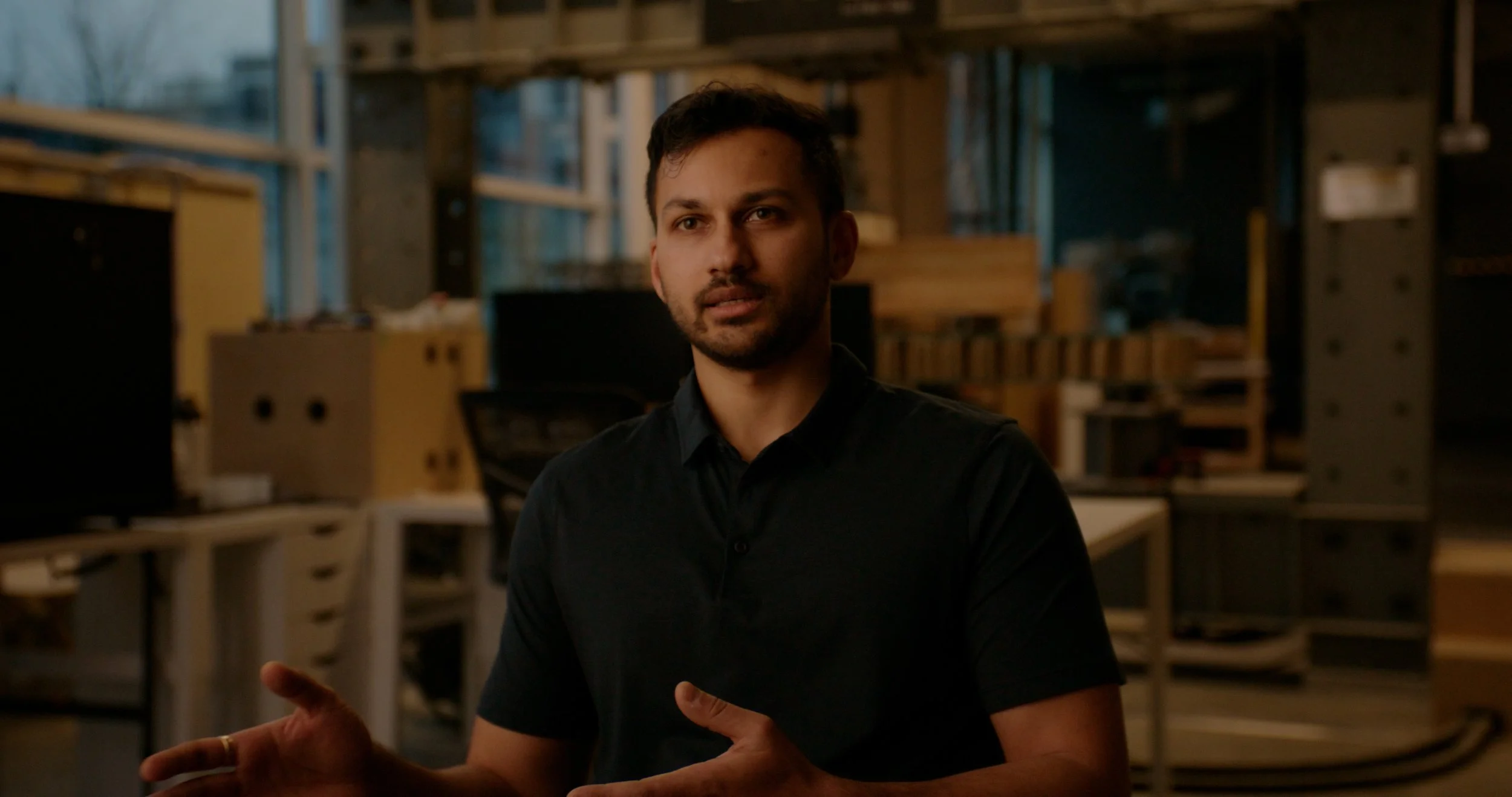 A man in a black shirt is talking, with a warehouse or workshop filled with shelves and boxes in the background.