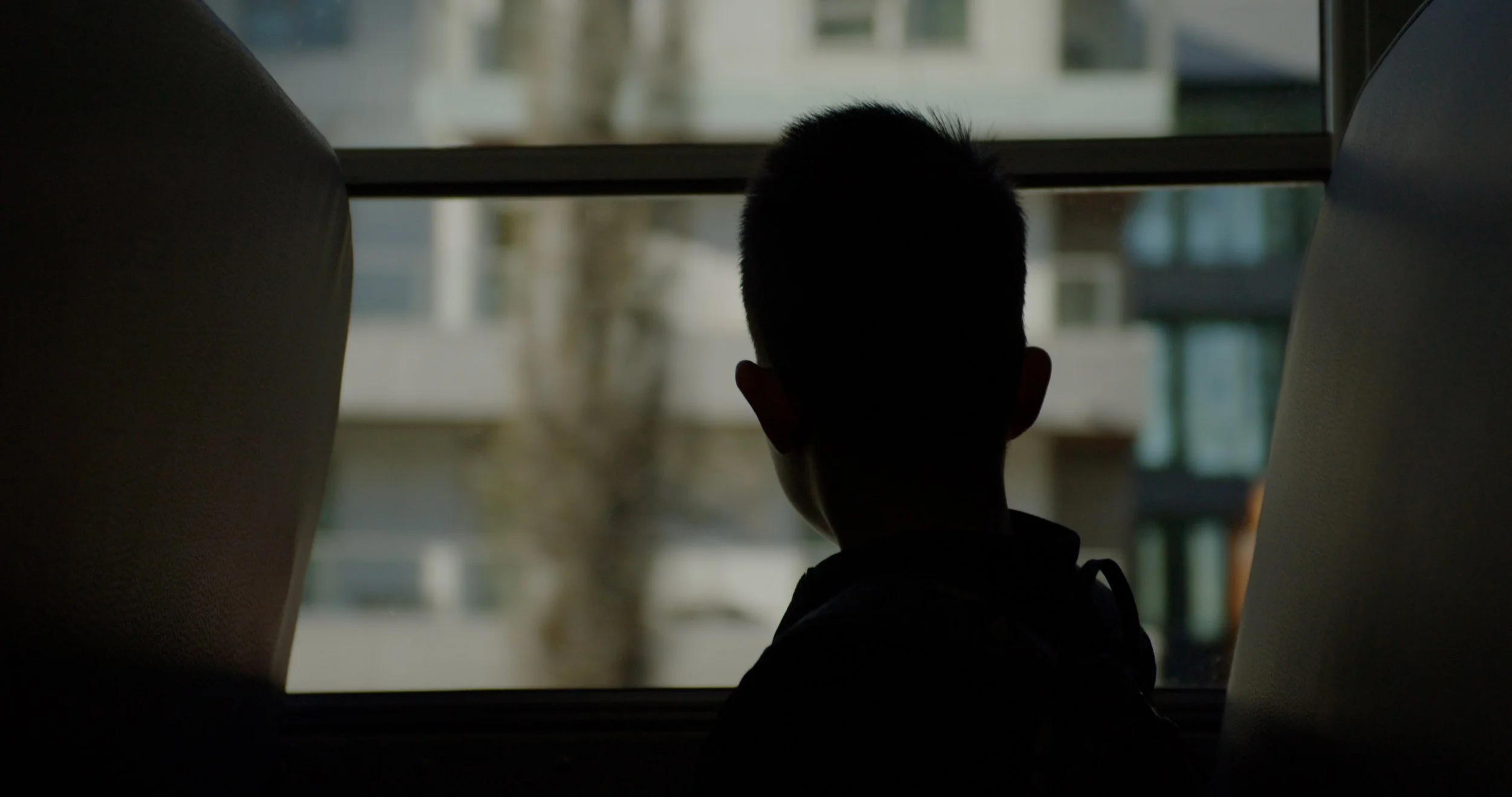 Silhouette of a young boy sitting on a bus or train, looking out the window at buildings outside.