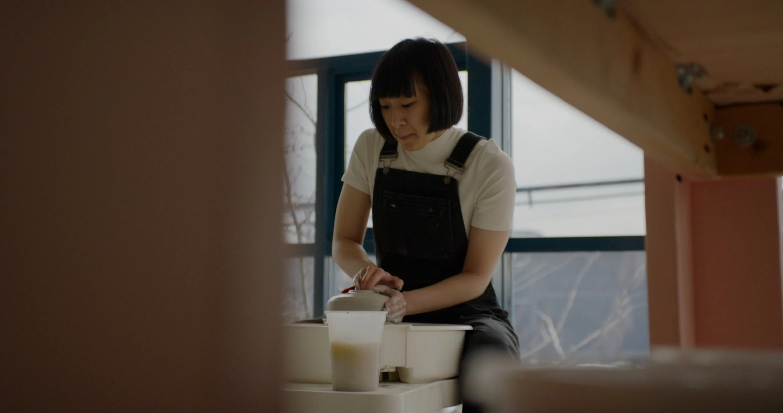A woman with short black hair, wearing a white t-shirt and black overalls, working on a pottery wheel in a studio with large windows in the background.