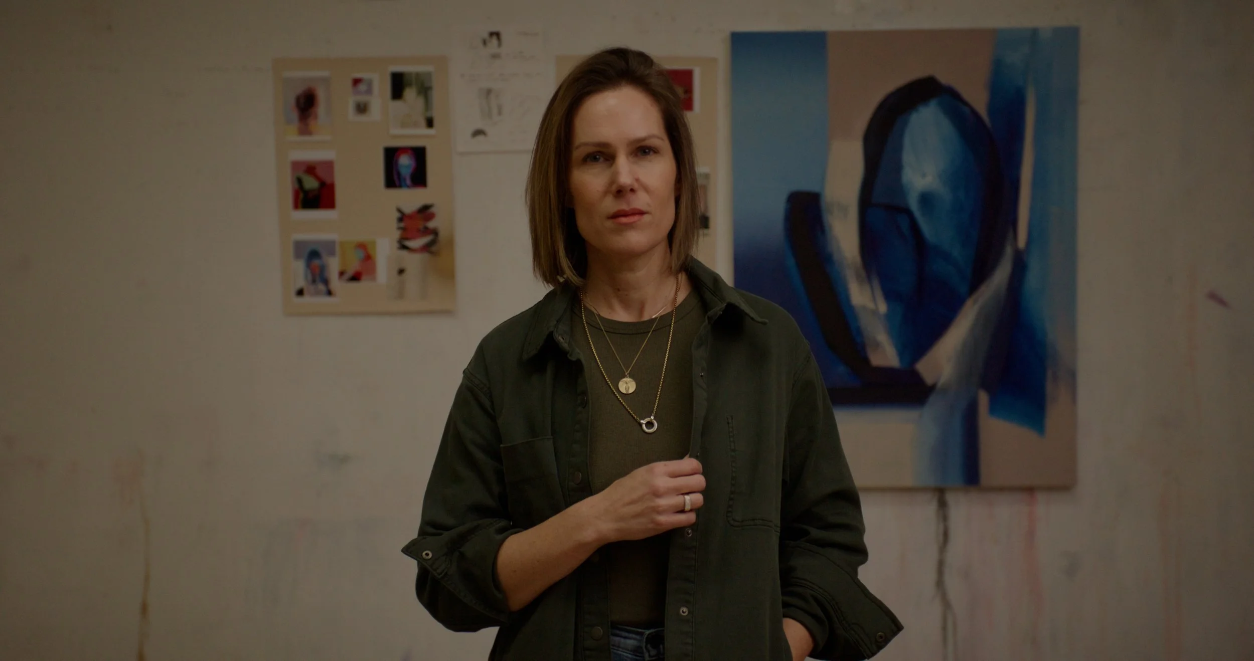 Woman with shoulder-length brown hair wearing a dark green shirt and layered necklaces, standing in front of abstract artwork on a wall.