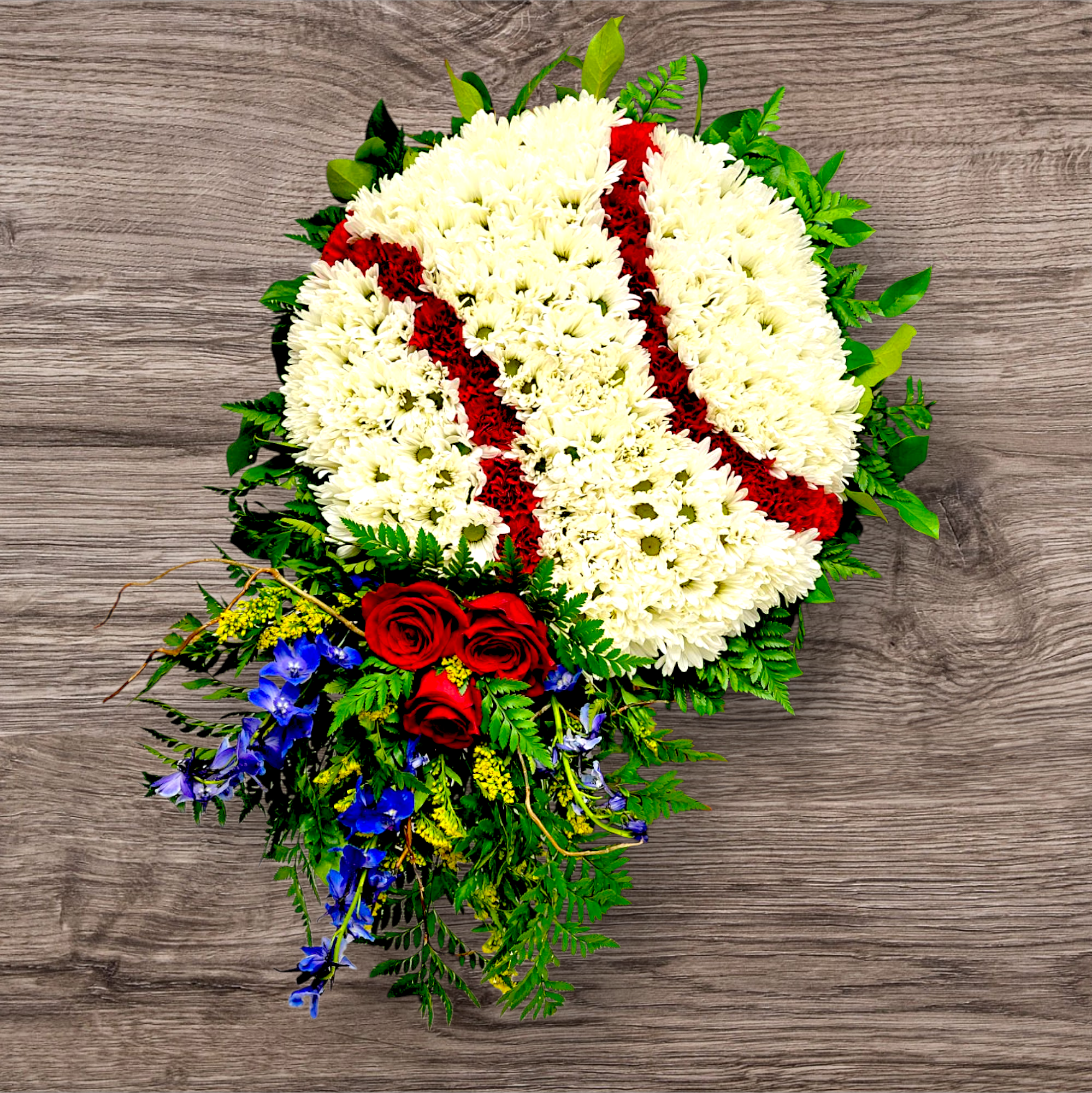A floral funeral wreath with white chrysanthemums, red roses, blue flowers, and green foliage on a wooden surface shaped as a baseball for a personal touch.