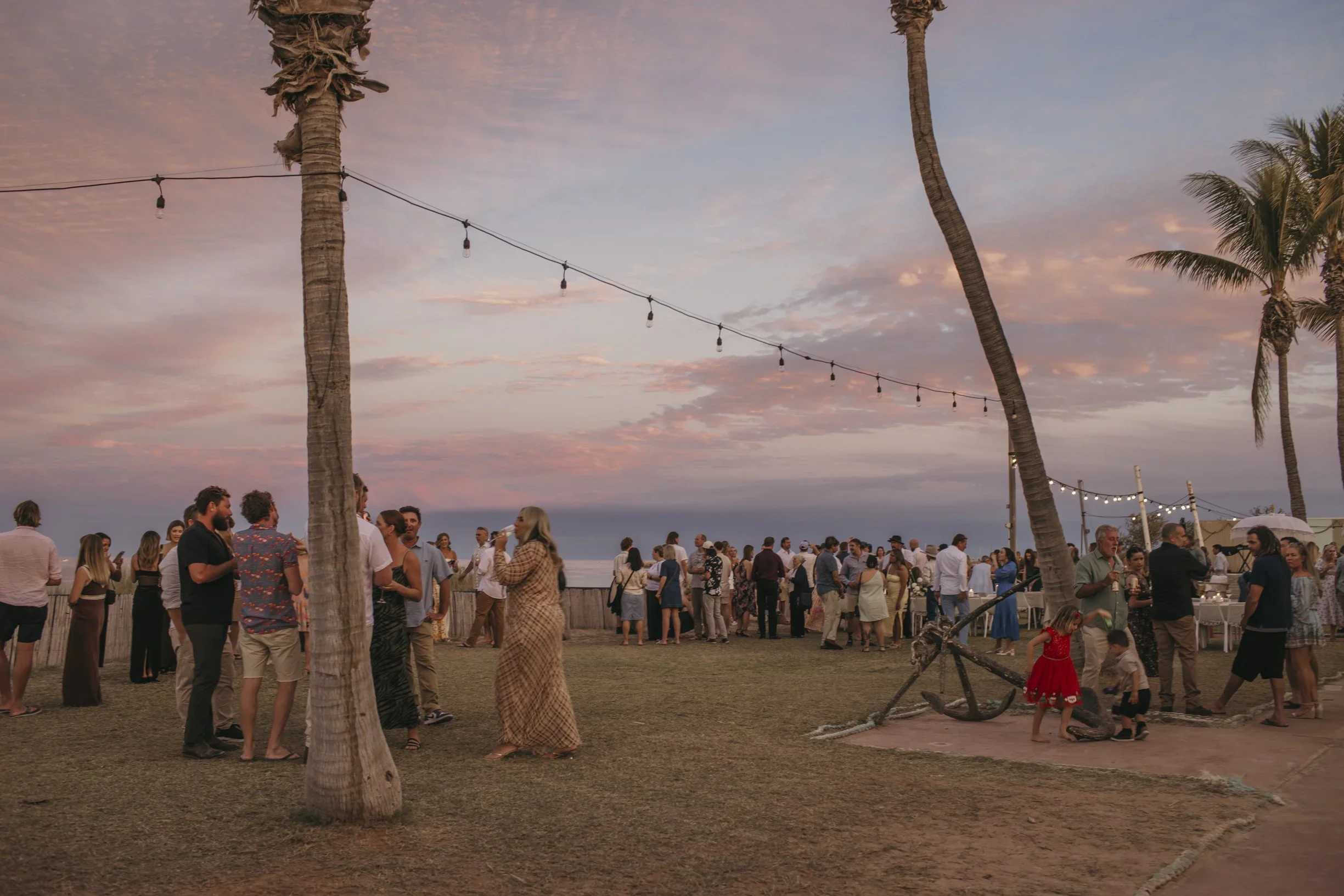 A crowd gathers around palm trees at sunset during a beach wedding in Exmouth, creating a romantic atmosphere.