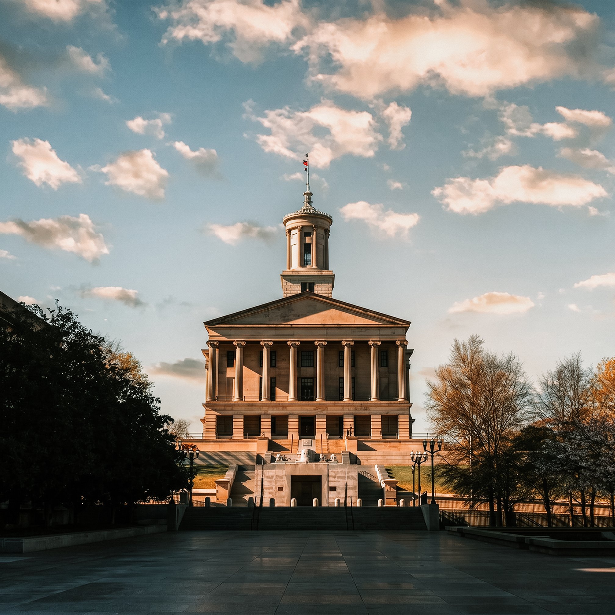 Tennessee state capitol with columns and a round tower with a clock and a Tennessee flag on top, set against a sky with scattered clouds during sunset, with trees and stairs in the foreground.