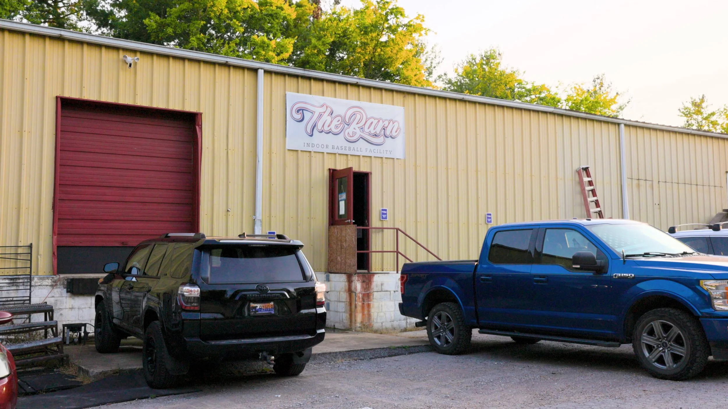 Outside view of a yellow metal building labeled 'The Barn Indoor Baseball Facility,' with several parked cars in front and trees in the background.