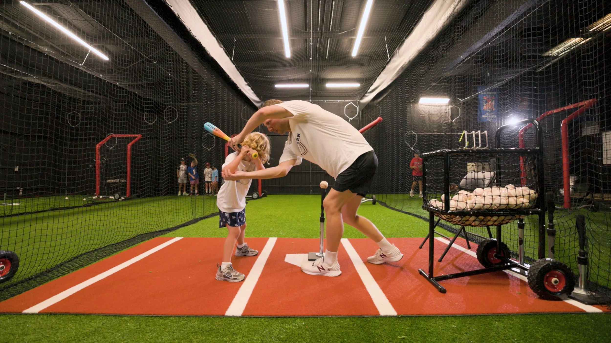 A man coaching a young boy in a batting stance inside a batting cage at an indoor baseball facility, with other children in the background.