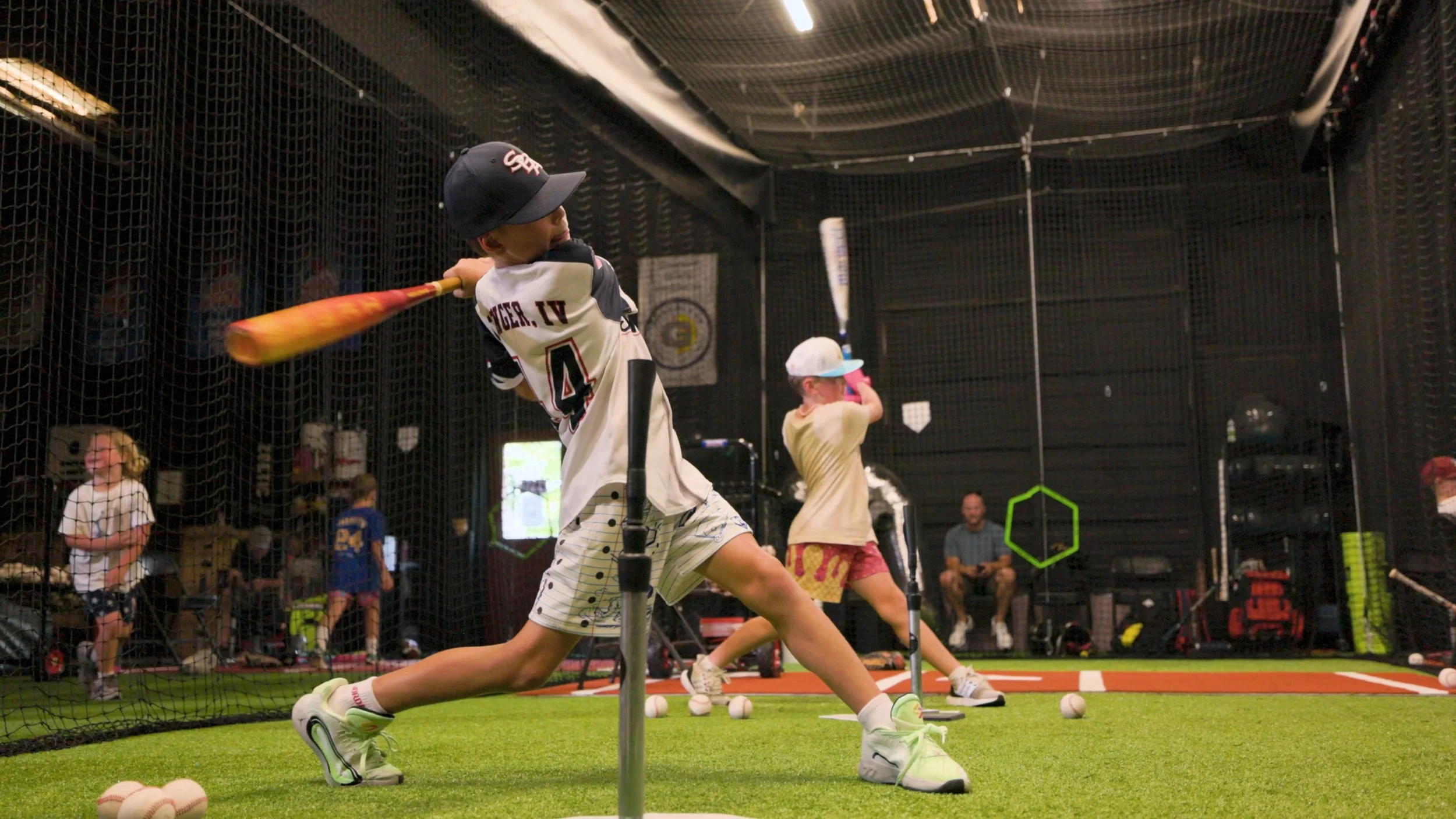 Young boy in sports attire practicing baseball batting at indoor batting cage, with other kids and adults in the background.