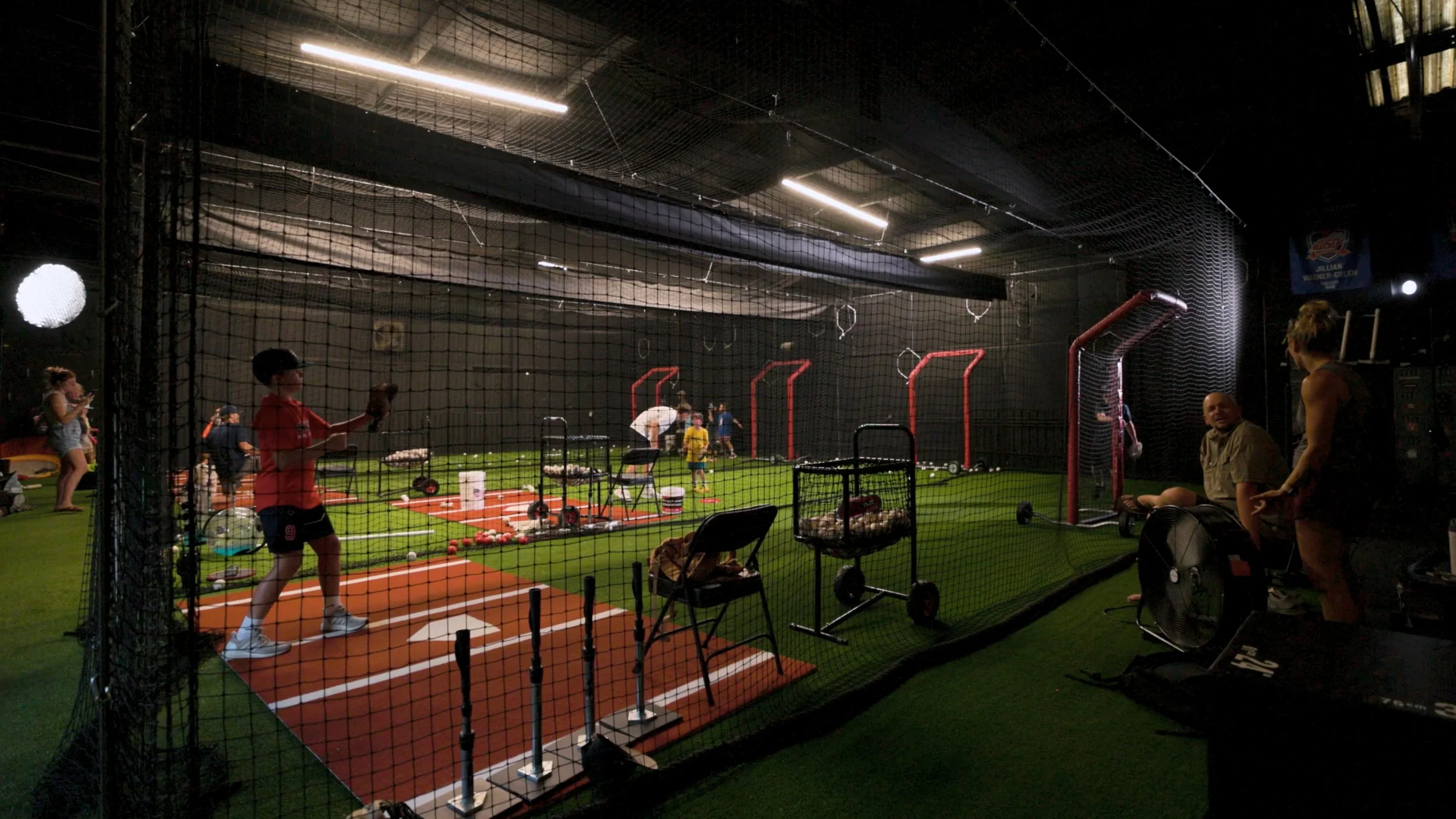Indoor baseball training facility with children practicing batting and throwing, netting around the area, and coaches supervising.