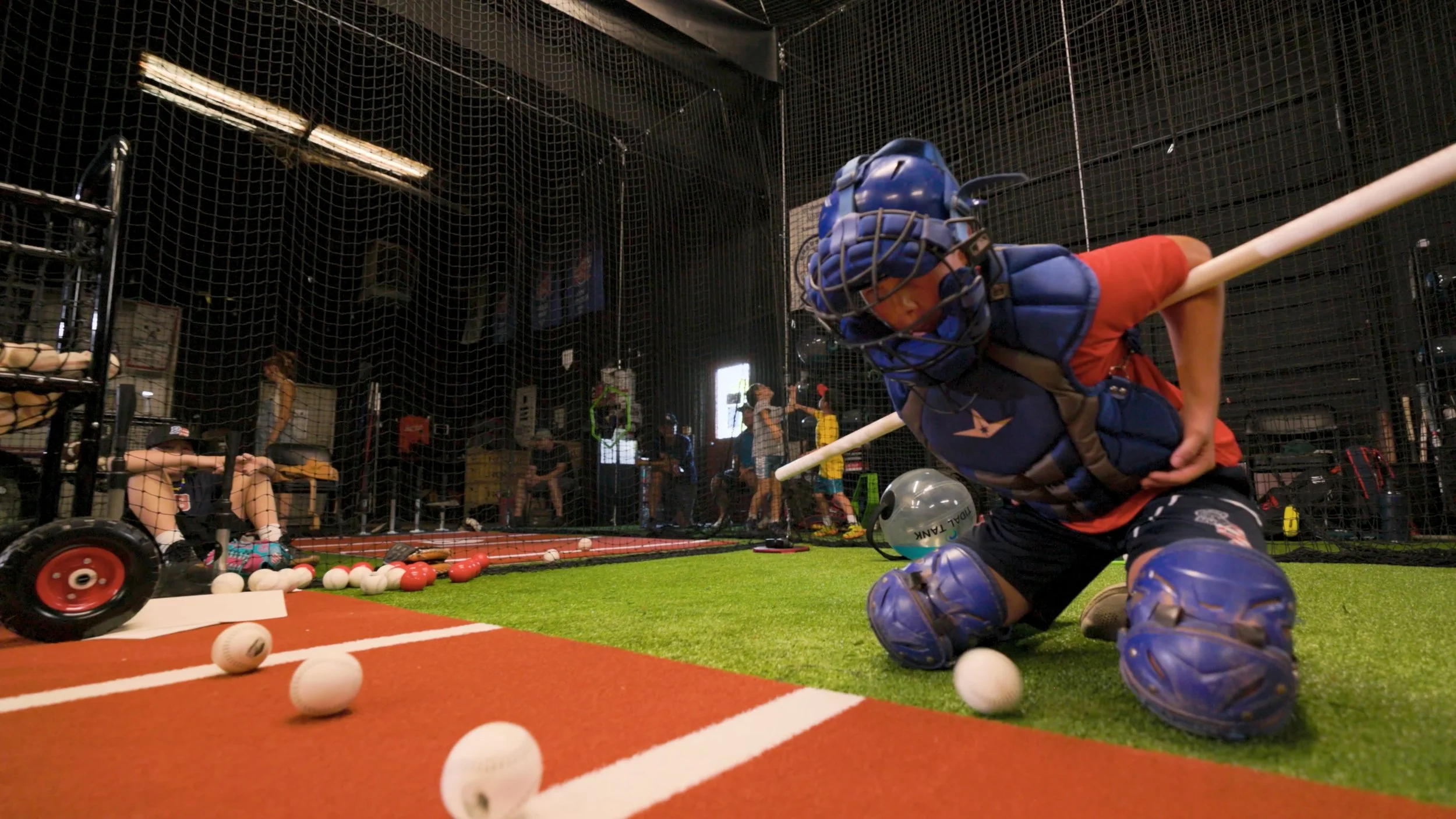 A young boy dressed in baseball catcher's gear practicing inside a batting cage, surrounded by various baseballs and softballs with other children and adults in the background.