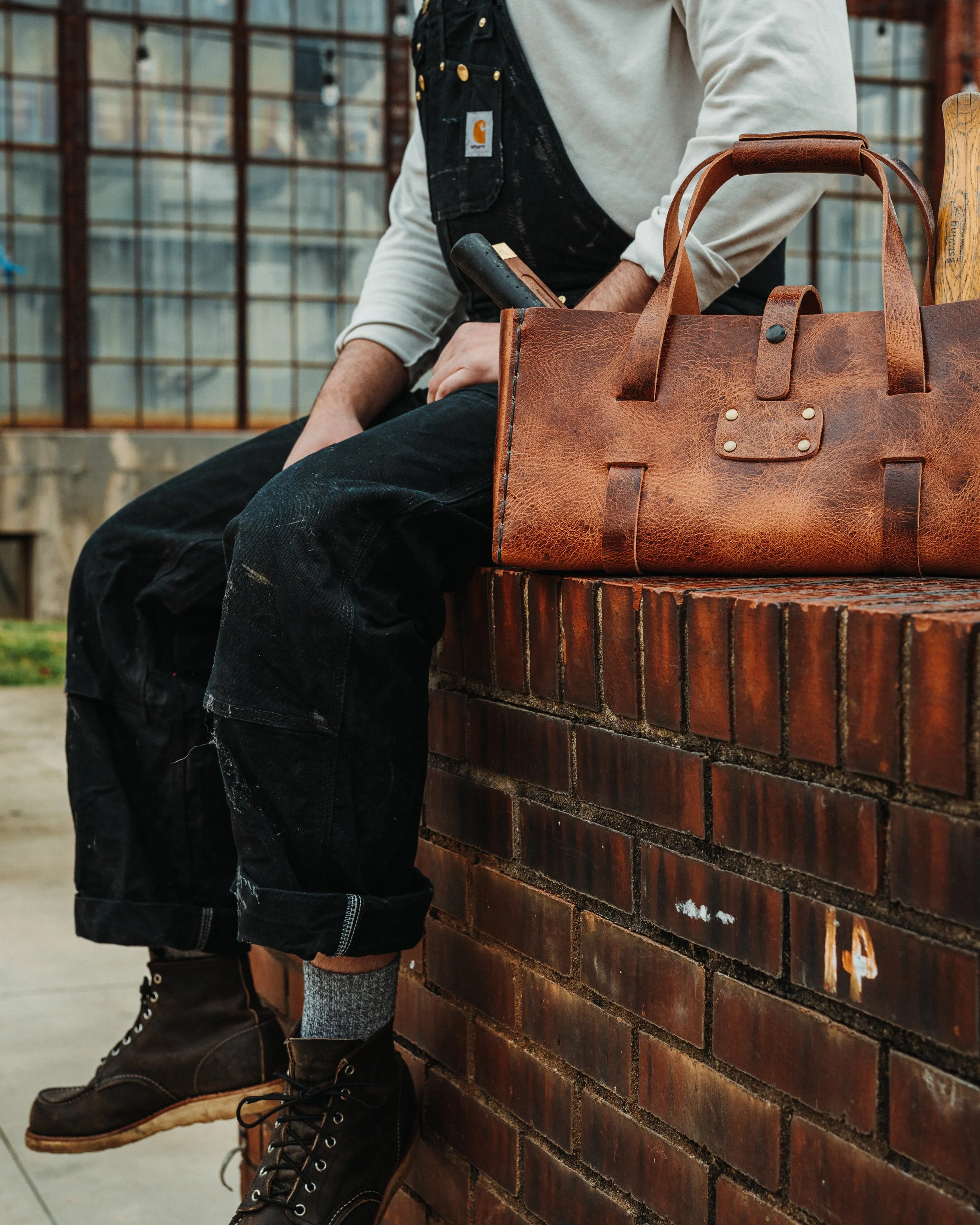 Close-up of a person sitting on a brick ledge, wearing black work boots, black shorts, a gray sweater, and a black overall with a Carhartt logo. They are holding a hammer behind a large brown leather bag with handles and a snap closure, and a small tool is sticking out of the bag. The background includes a building with glass windows.