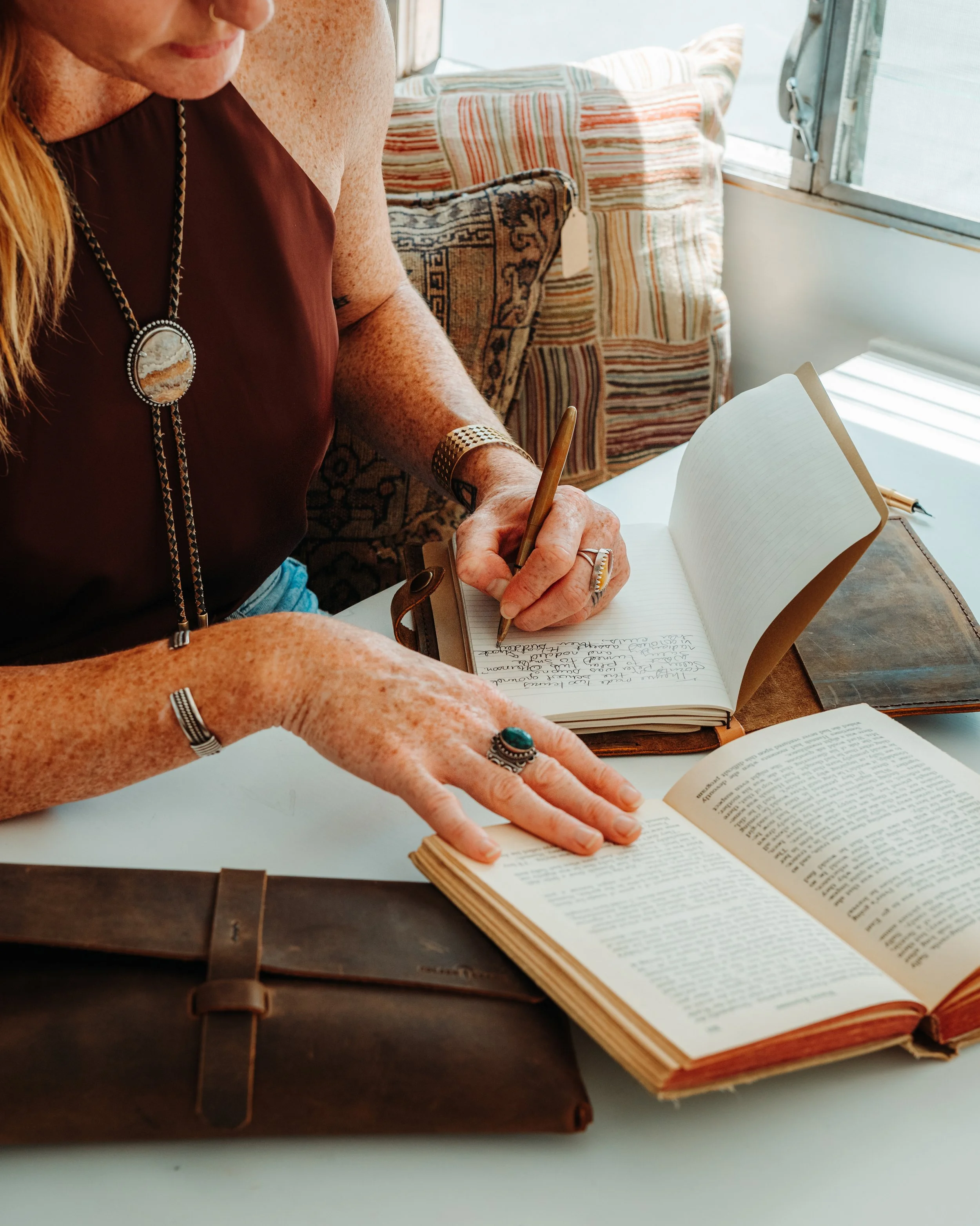 A woman with red hair and freckles is sitting at a table, writing in a notebook. She is wearing a brown sleeveless top, a large pendant necklace, a wide bracelet, and rings. There are open books in front of her and colorful, patterned pillows and an open window are in the background.