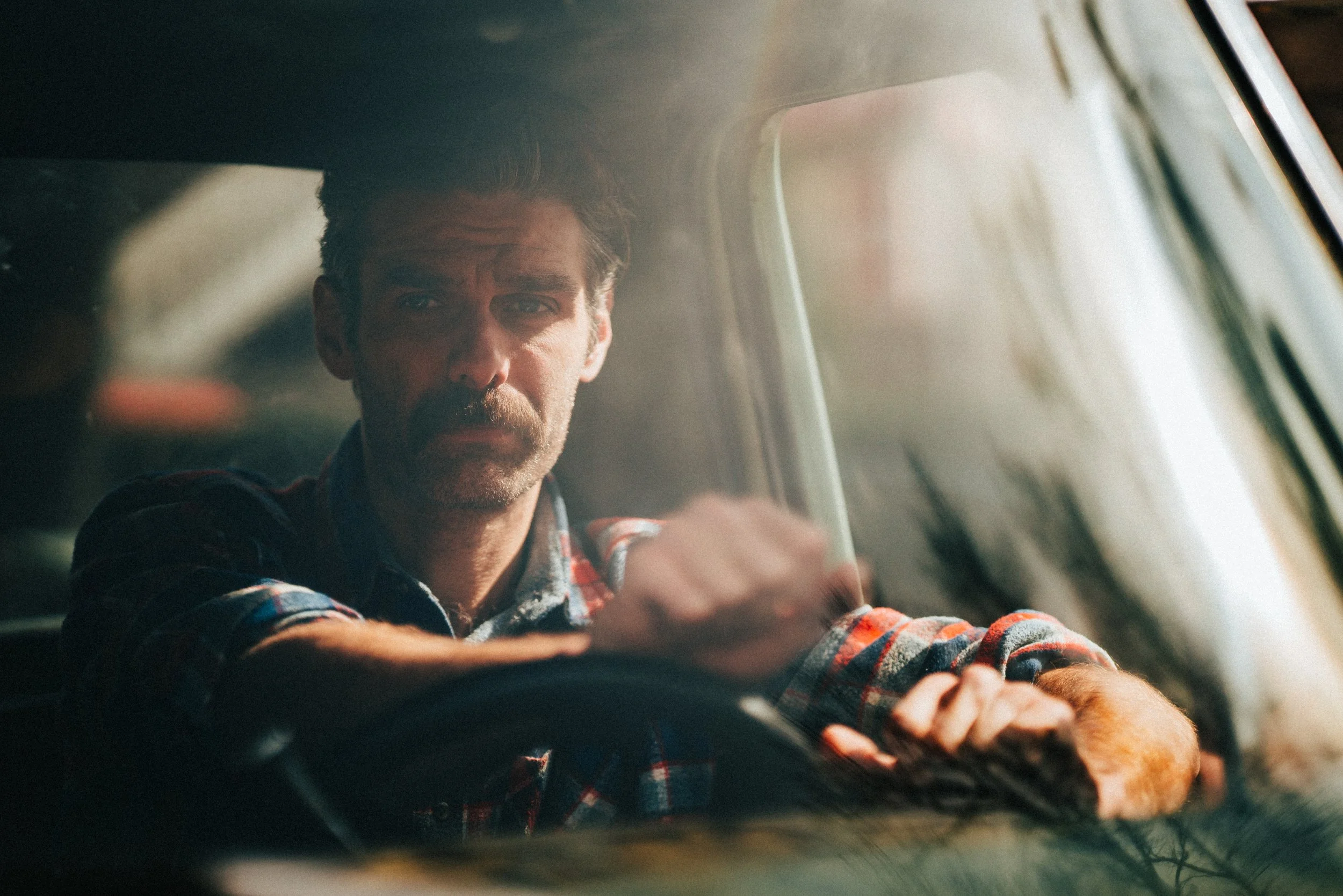 Man with a beard and mustache wearing a red plaid shirt, sitting inside a vehicle, with sunlight shining through the window.