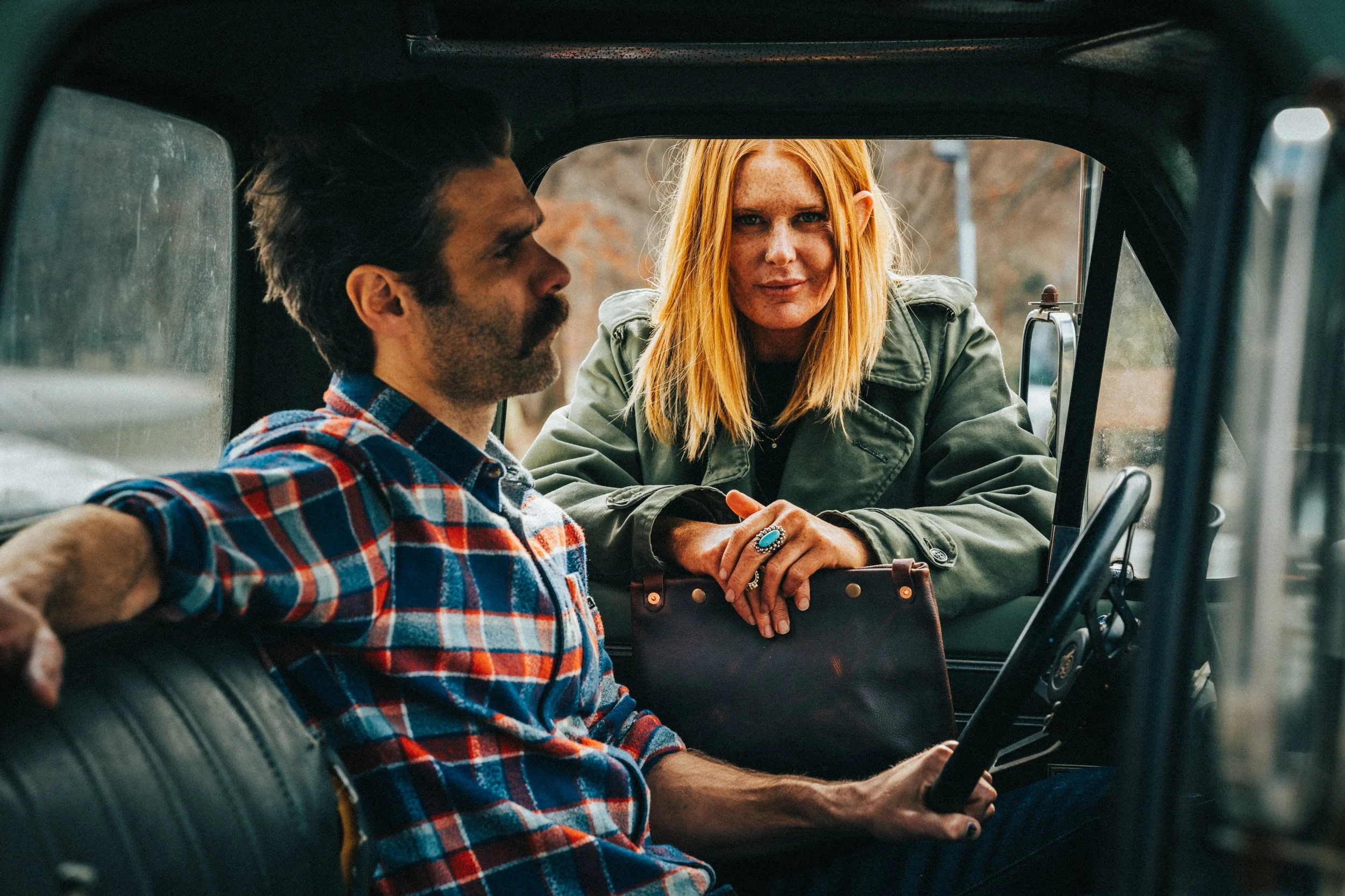 A man with dark hair and a beard wearing a red and blue plaid shirt and a woman with long red hair, freckles, and green jacket sitting inside a vehicle, with the woman leaning in the open vehicle door.