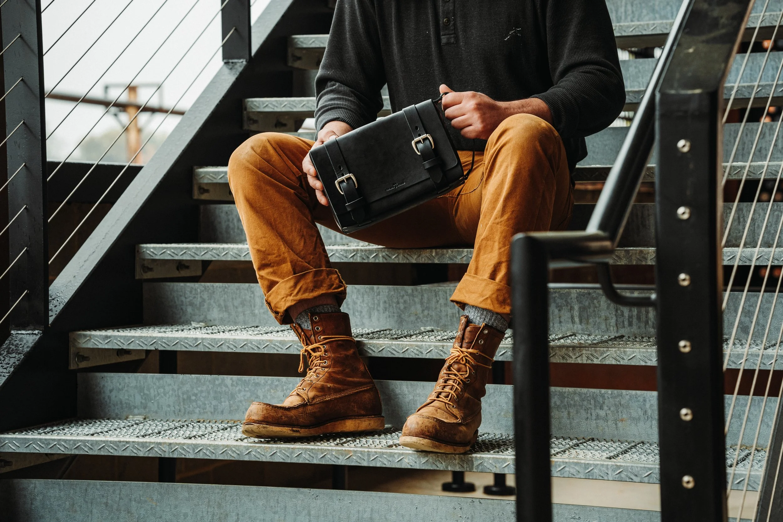 Person sitting on metal stairs holding a black bag, wearing brown pants and brown boots.