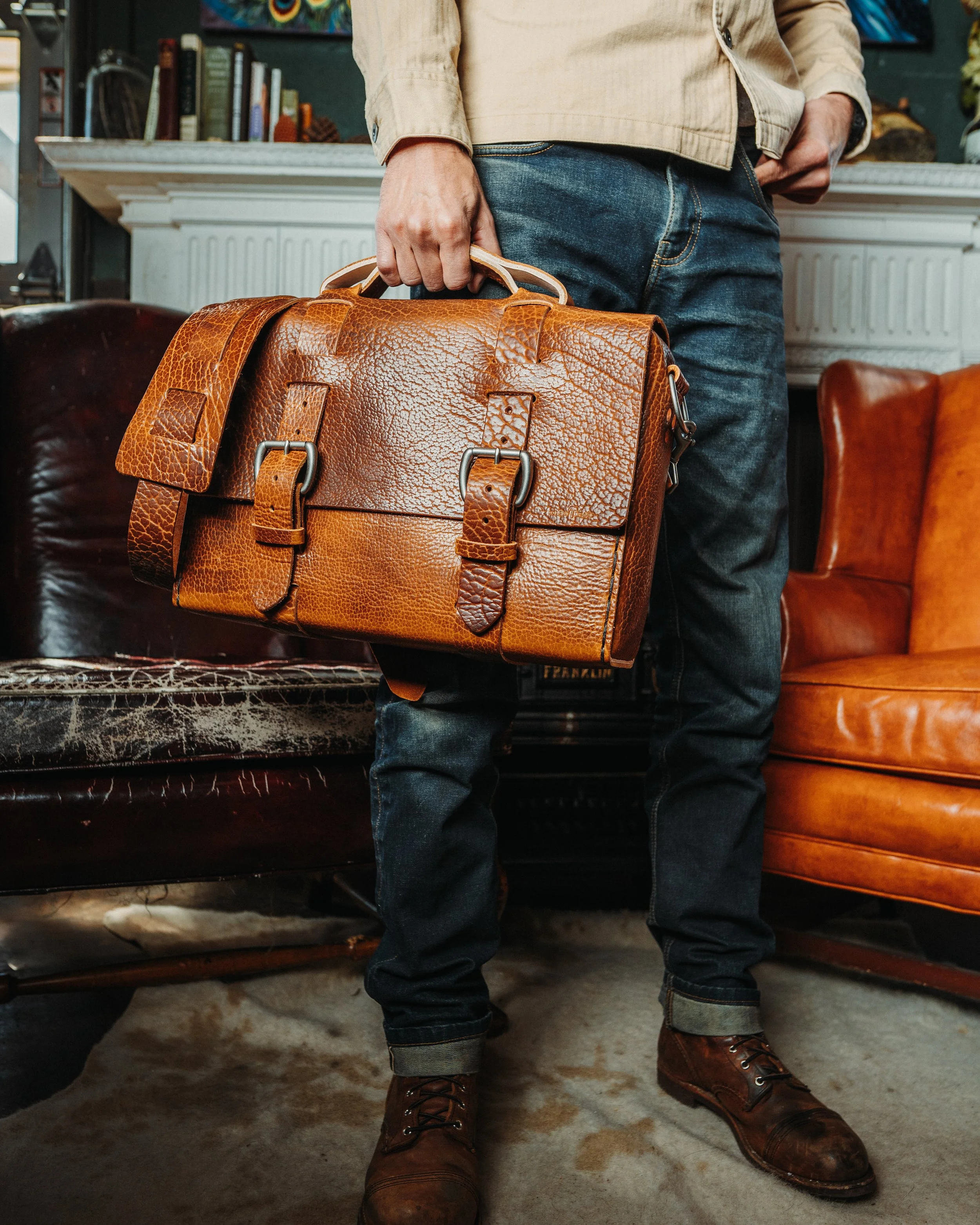 Person standing indoors holding a leather briefcase, wearing a beige jacket, blue jeans, and brown boots, with a background of bookshelves and leather chairs.