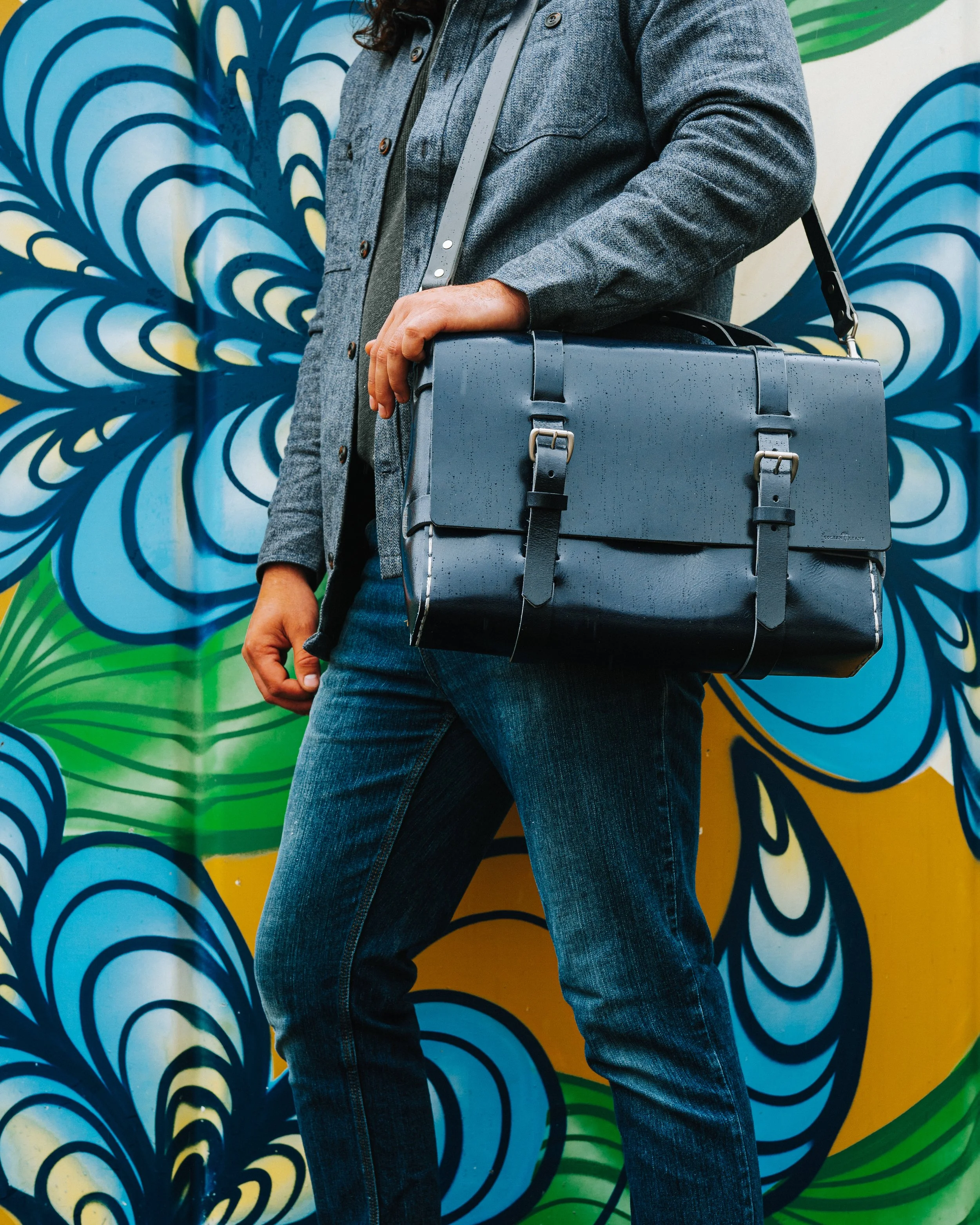 Person wearing a gray denim jacket and jeans holding a dark gray briefcase, standing in front of a vibrant, colorful mural with swirling blue, yellow, green, and black patterns.
