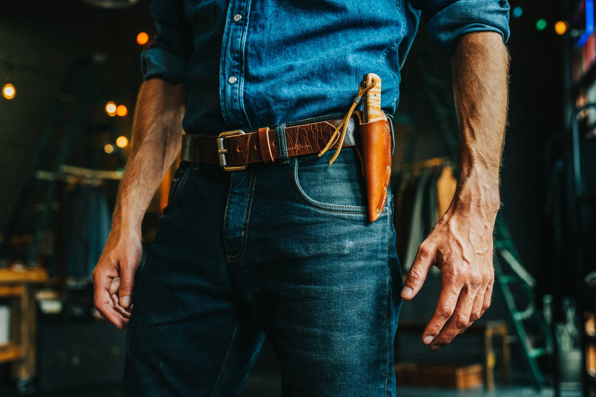 Close-up of a man wearing a denim shirt and jeans, with a leather belt that holds a knife with a wooden handle and a small leather pouch.