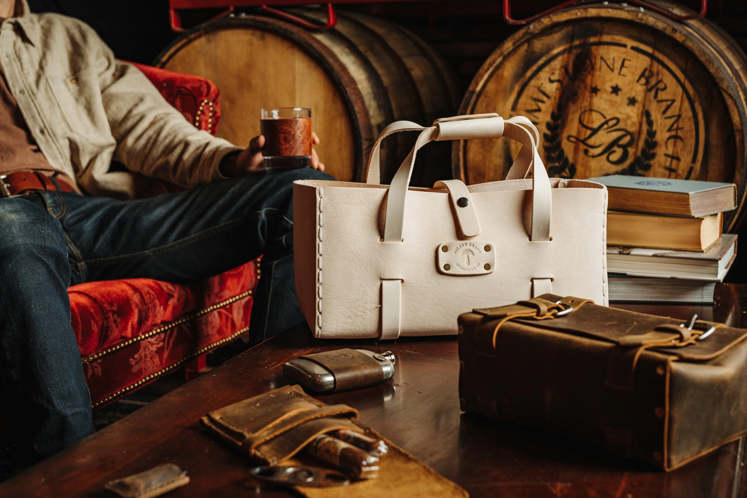 Person sitting on a red vintage couch holding a glass of dark beverage, surrounded by leather wallets, a bag, and a book with wooden barrels in the background.