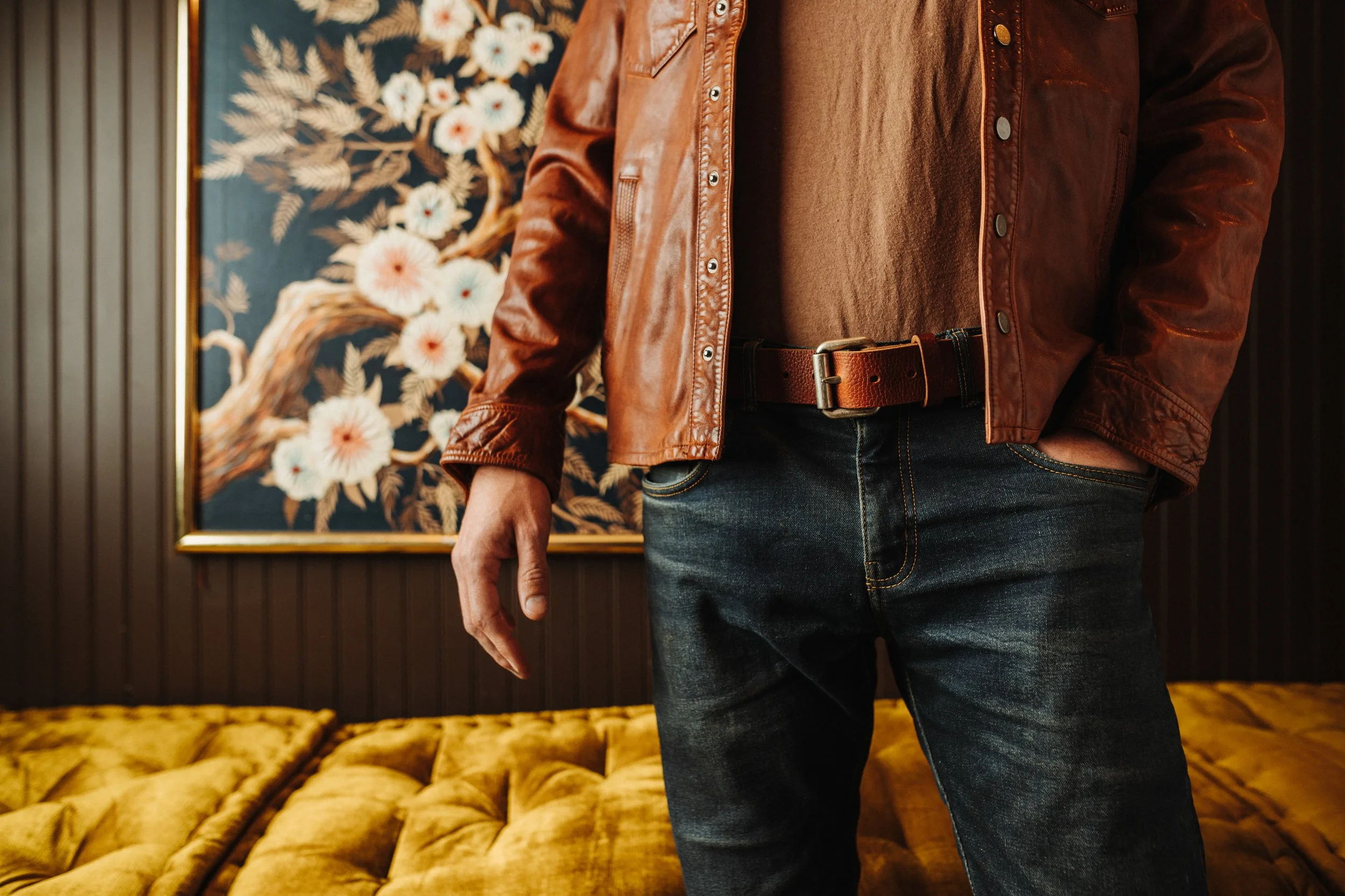 Close-up of a person wearing a brown leather jacket, a brown shirt, and dark jeans, standing in a room with a gold tufted sofa, floral artwork, and wood-paneled walls.