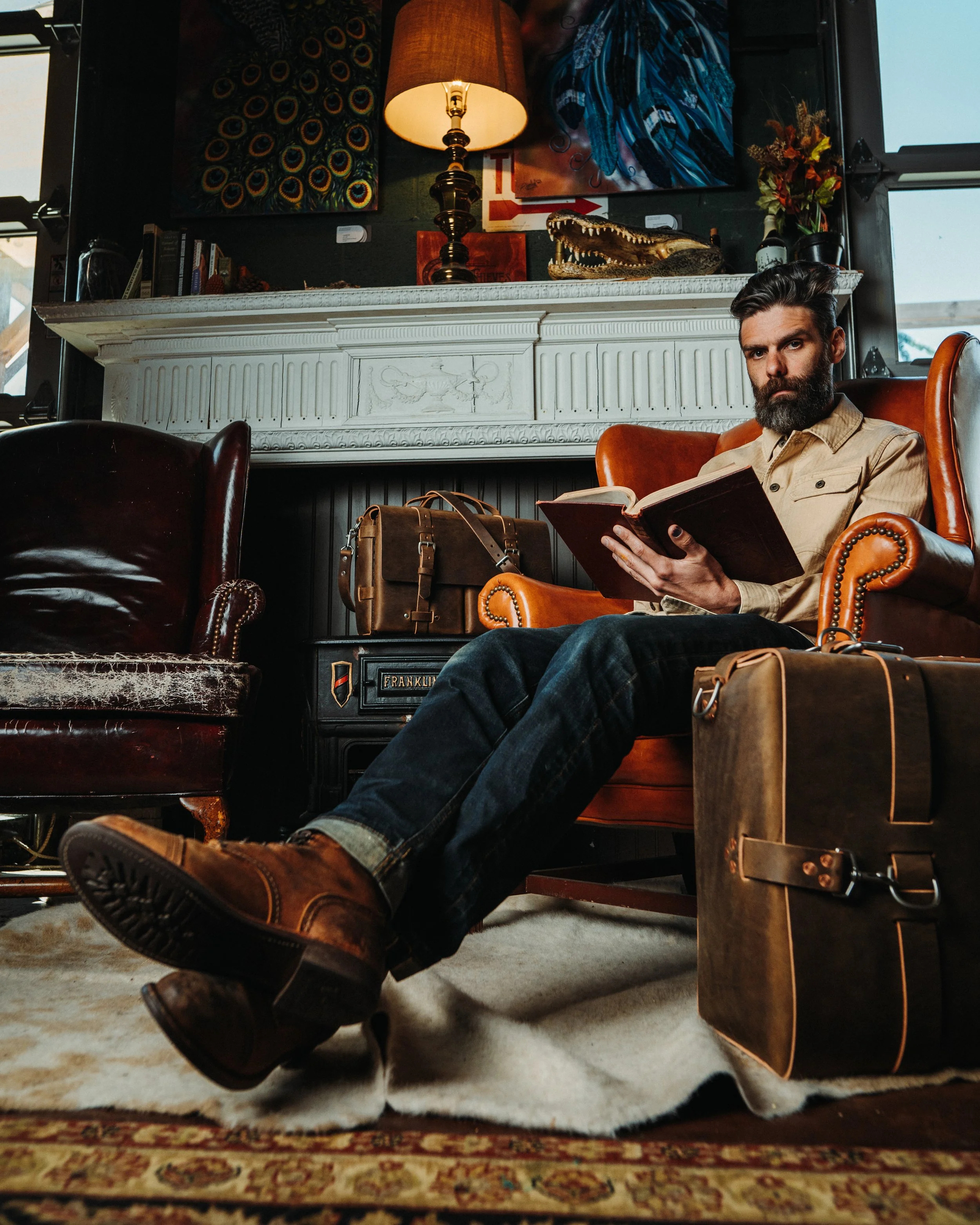 A man with a beard and dark hair sitting in a leather armchair reading a book in a cozy living room with a fireplace mantel decorated with artwork, a lamp, a crocodile model, and various decorative items.