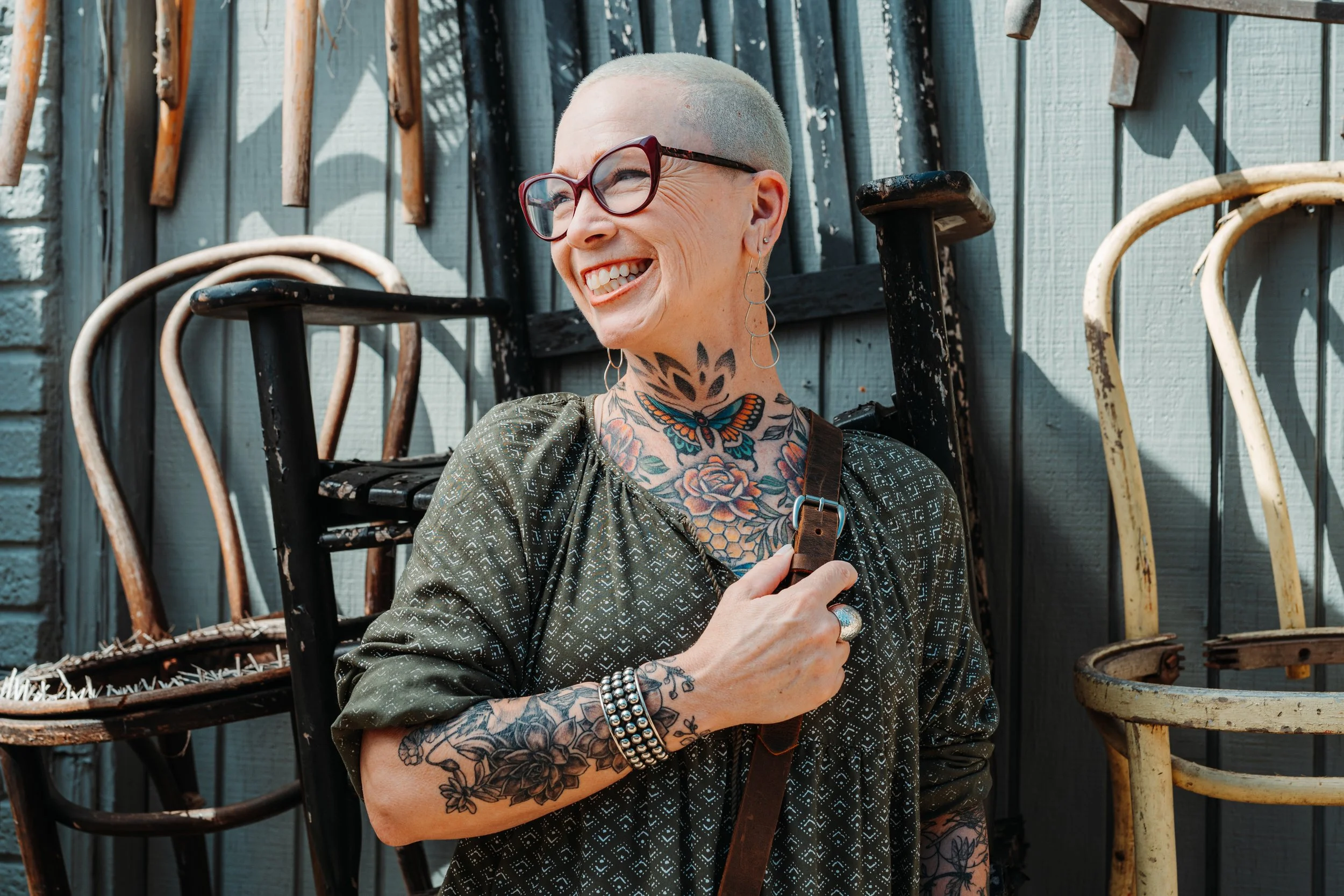 Smiling woman with glasses and tattoos, standing outdoors in front of a wooden fence and vintage chairs, holding a leather strap.