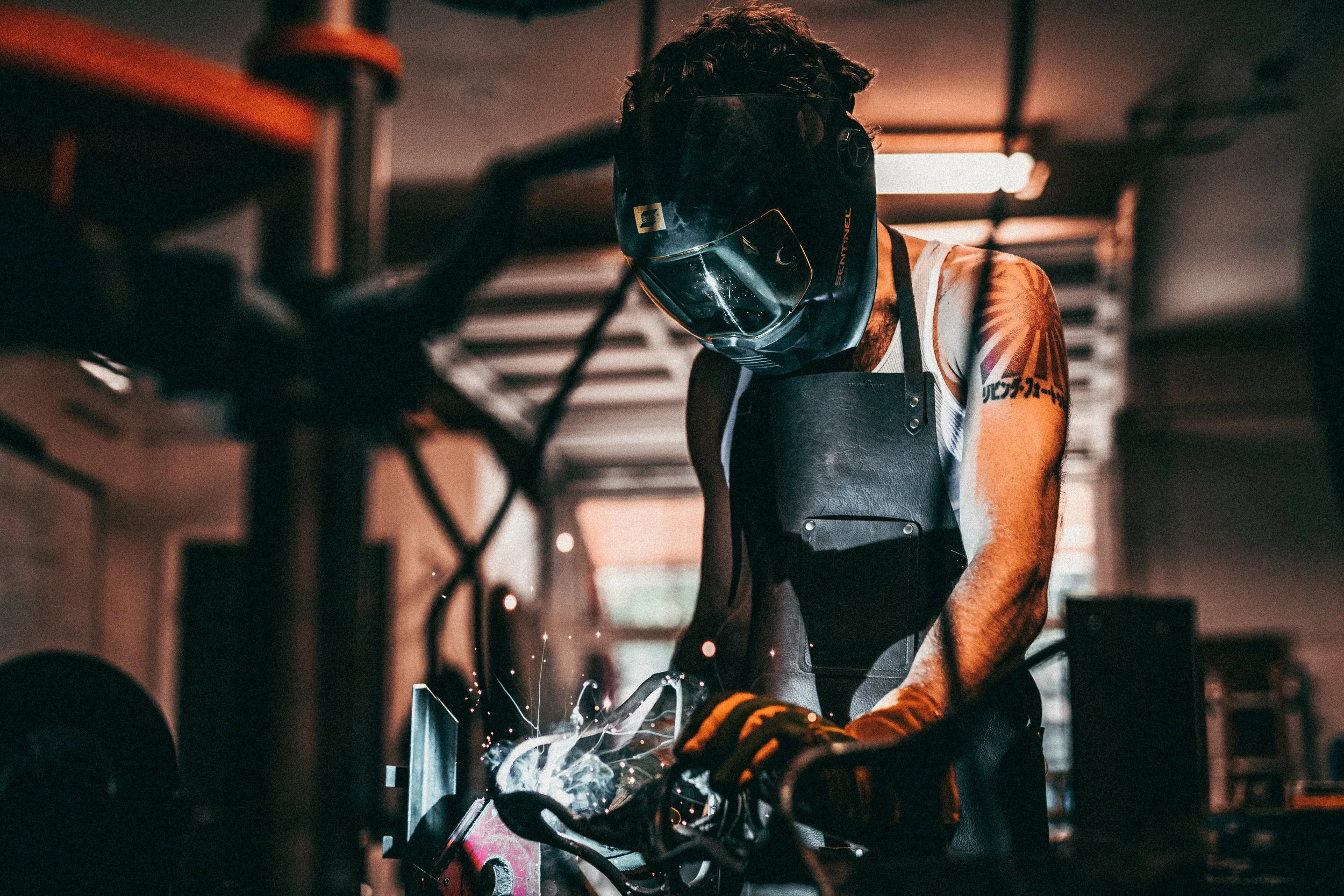 A person wearing a welding helmet and protective gear welding metal, with sparks flying around, in a workshop or garage setting.