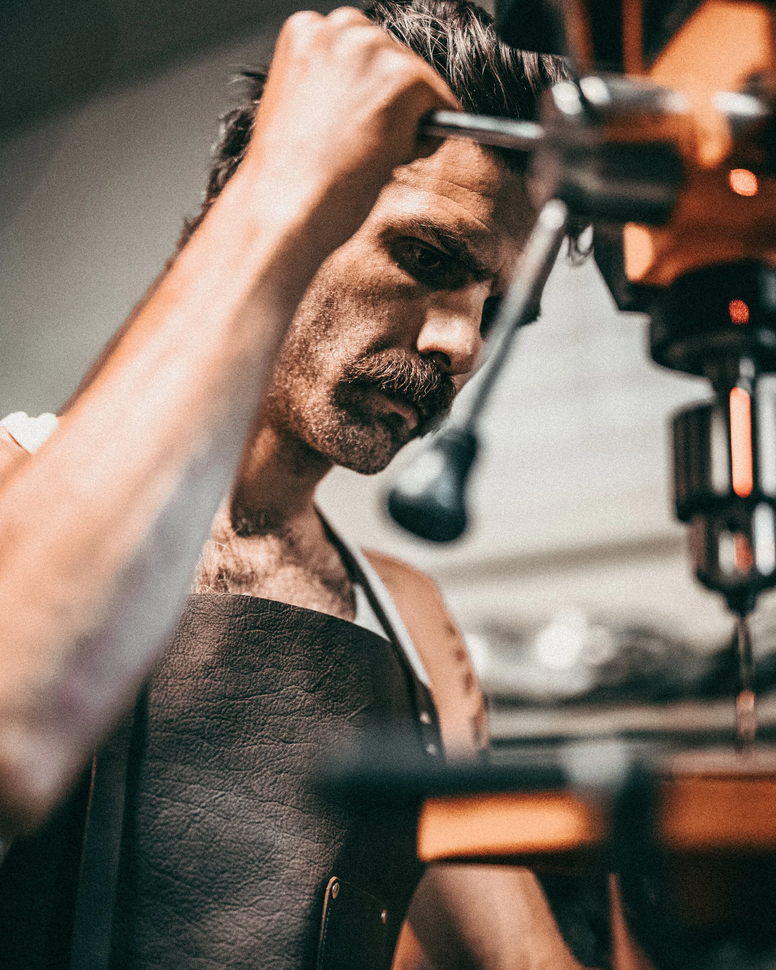 Close-up of a man with a beard and mustache, wearing a leather apron, lowering a drill press in a welding shop.