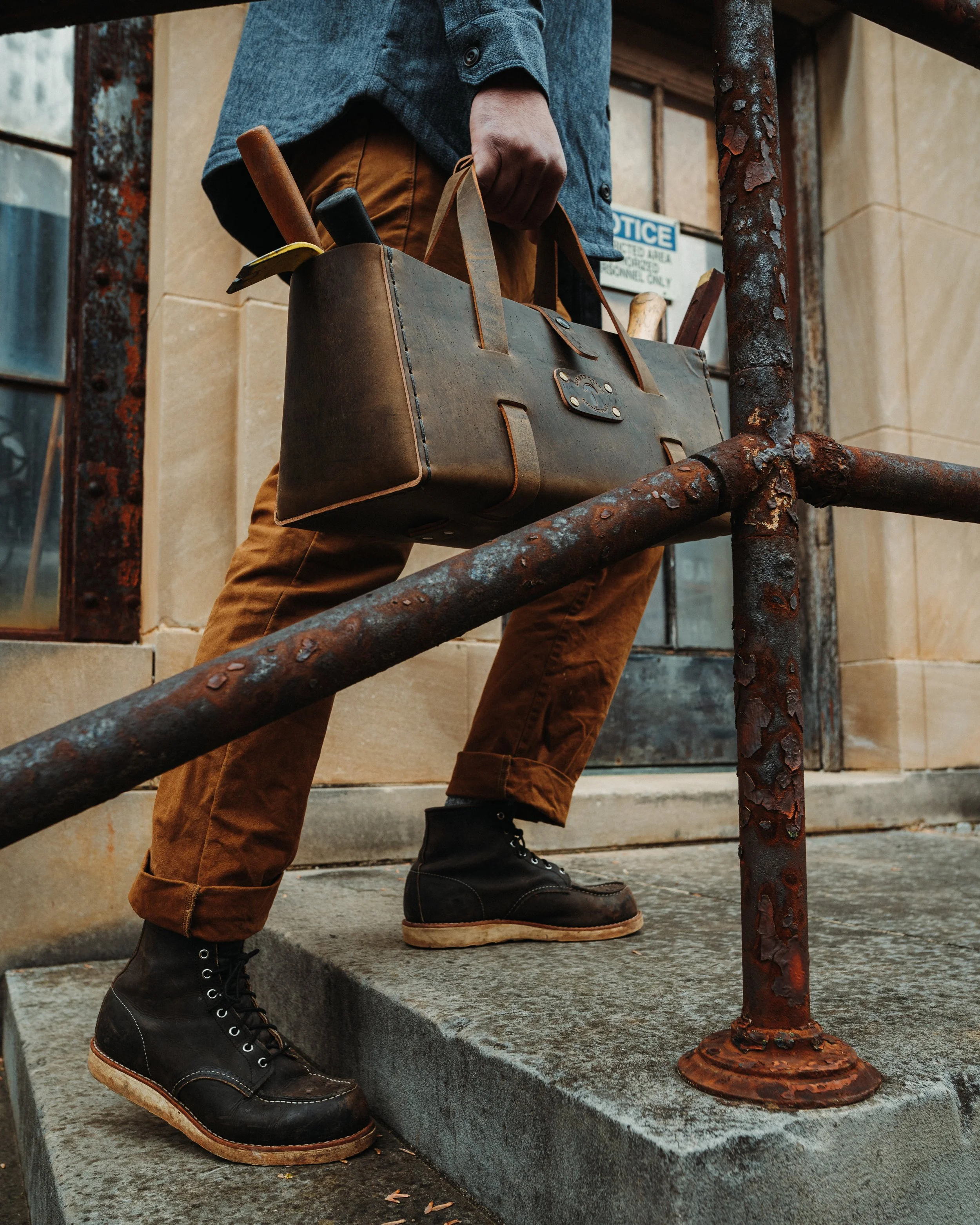 Person standing on a stone staircase holding a brown leather toolbox with a hammer and other tools inside. The person is wearing dark brown pants, a blue jacket, and black lace-up boots, and is near rusted metal railings and an old building.