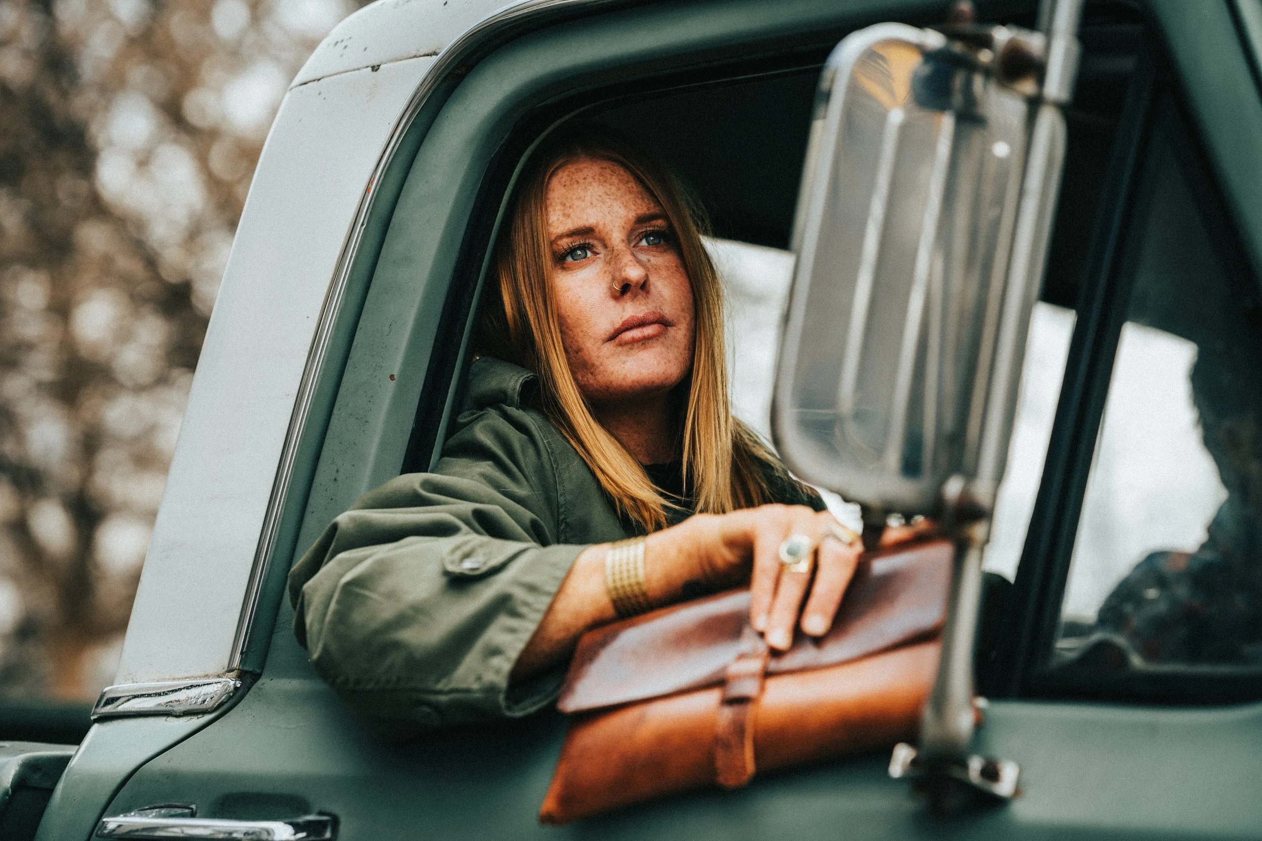 A woman with red hair and freckles looking out of the window of a vintage car, with her arm resting on the window frame. The car's exterior is a light color, and the window has a rounded shape. The background appears to be blurred outdoors.