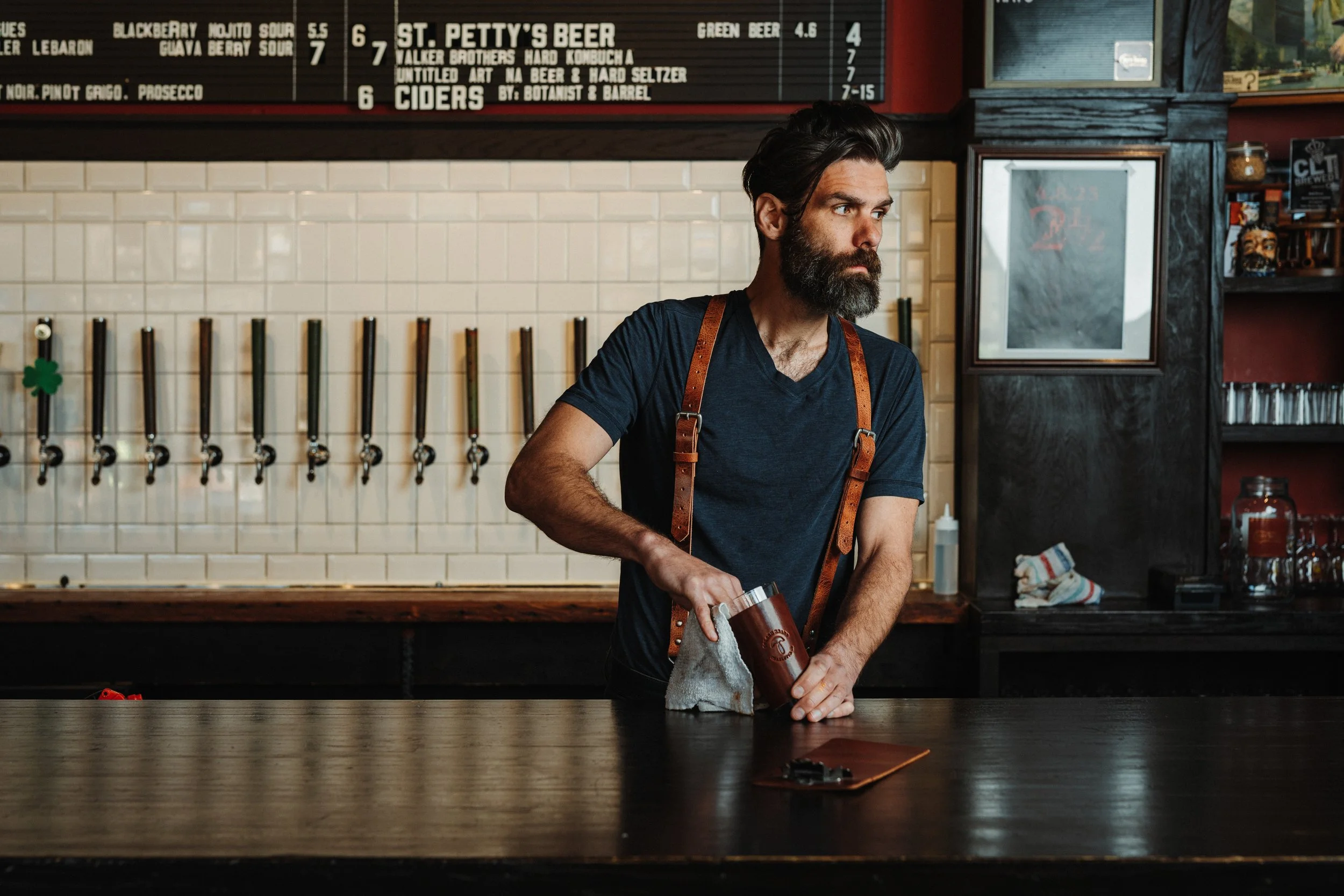 A bearded man with dark hair wearing a black t-shirt and a leather apron standing behind a bar counter in a restaurant or bar. He is holding a cloth and a drink container. The background features a blackboard menu with drink options, a tiled wall with beer taps, and some shelves with decorative items.