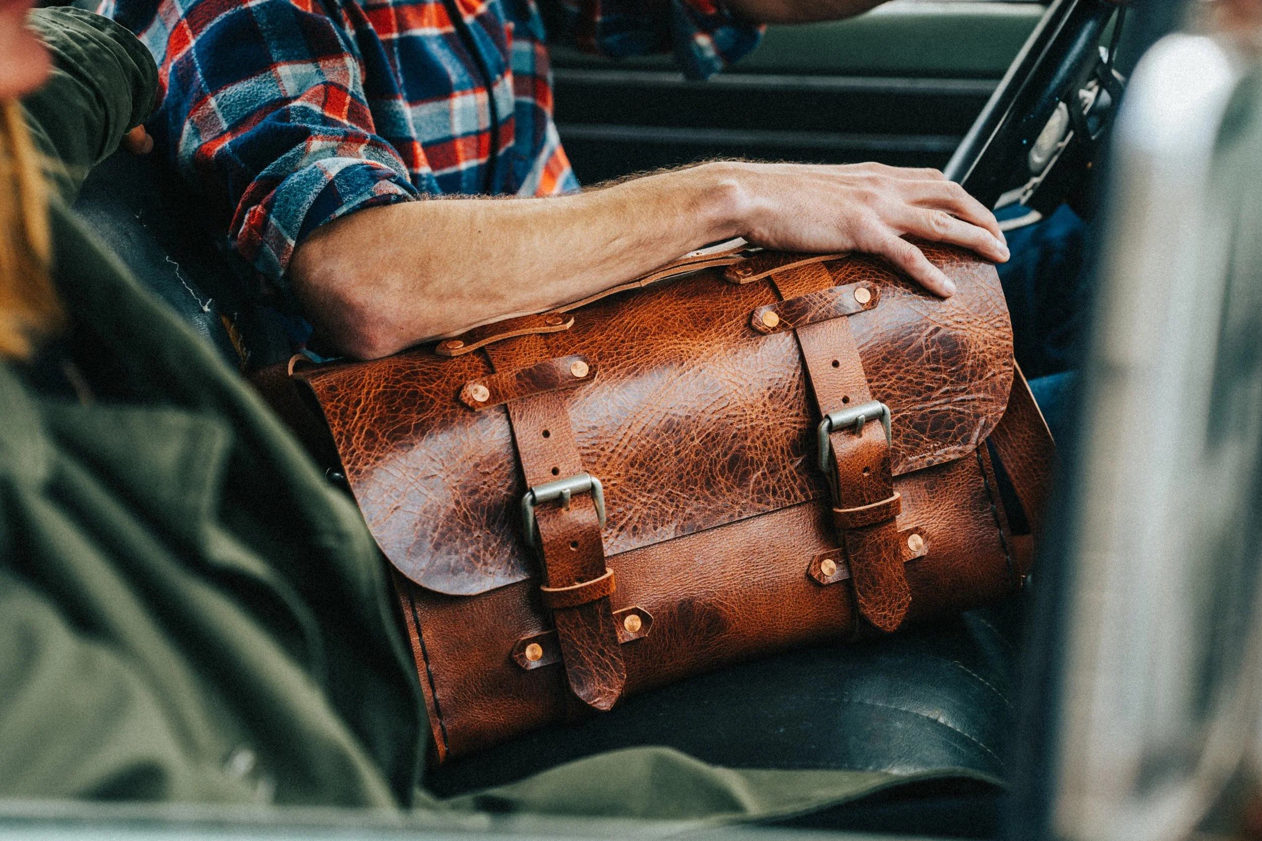 Close-up of a person's arm resting on a brown leather satchel inside a vehicle. The person is wearing a plaid shirt.