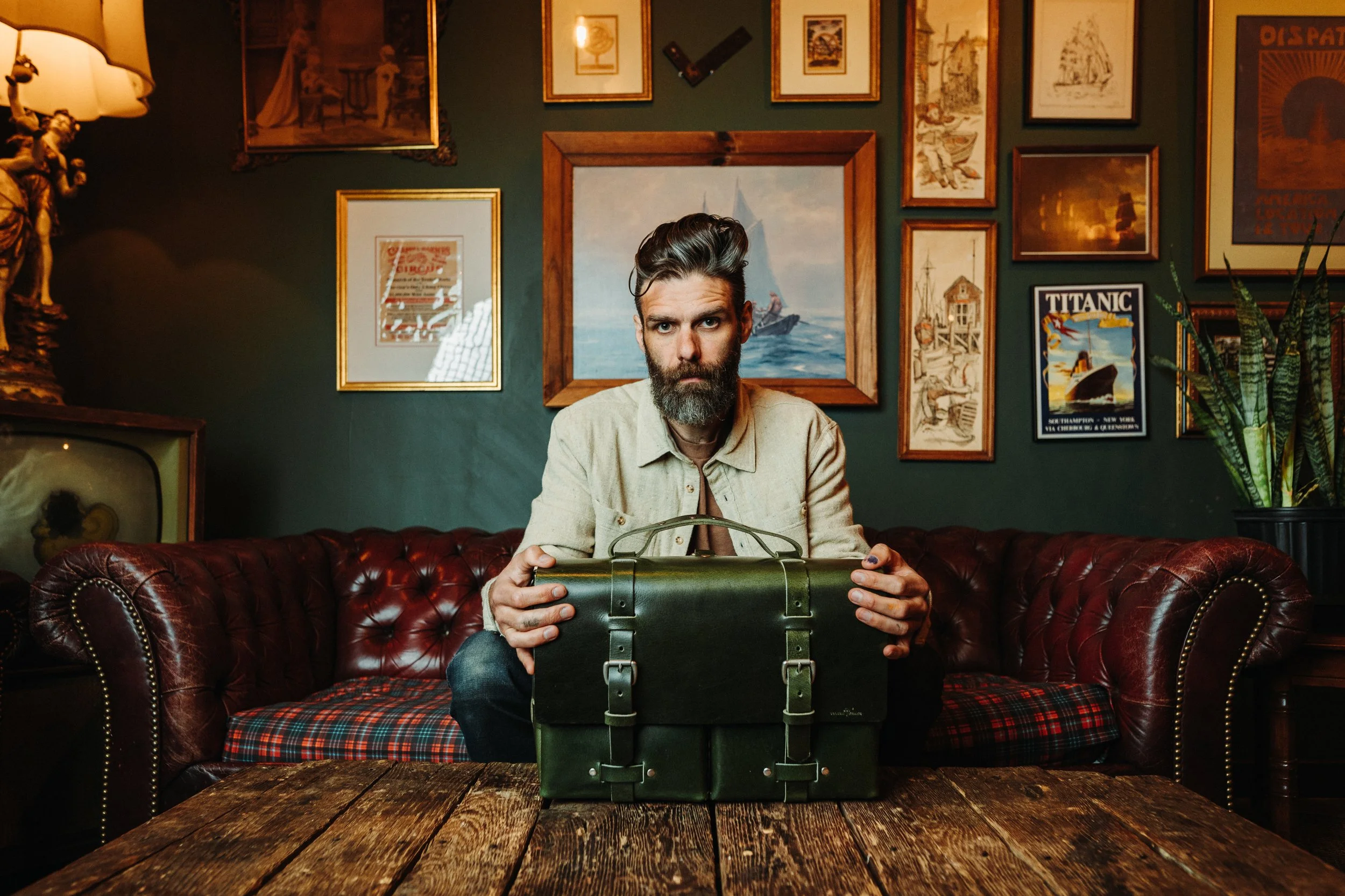 A man with a beard and styled hair sitting on a red leather sofa, holding a vintage green metal box, in a room decorated with various framed pictures and artwork on the wall.