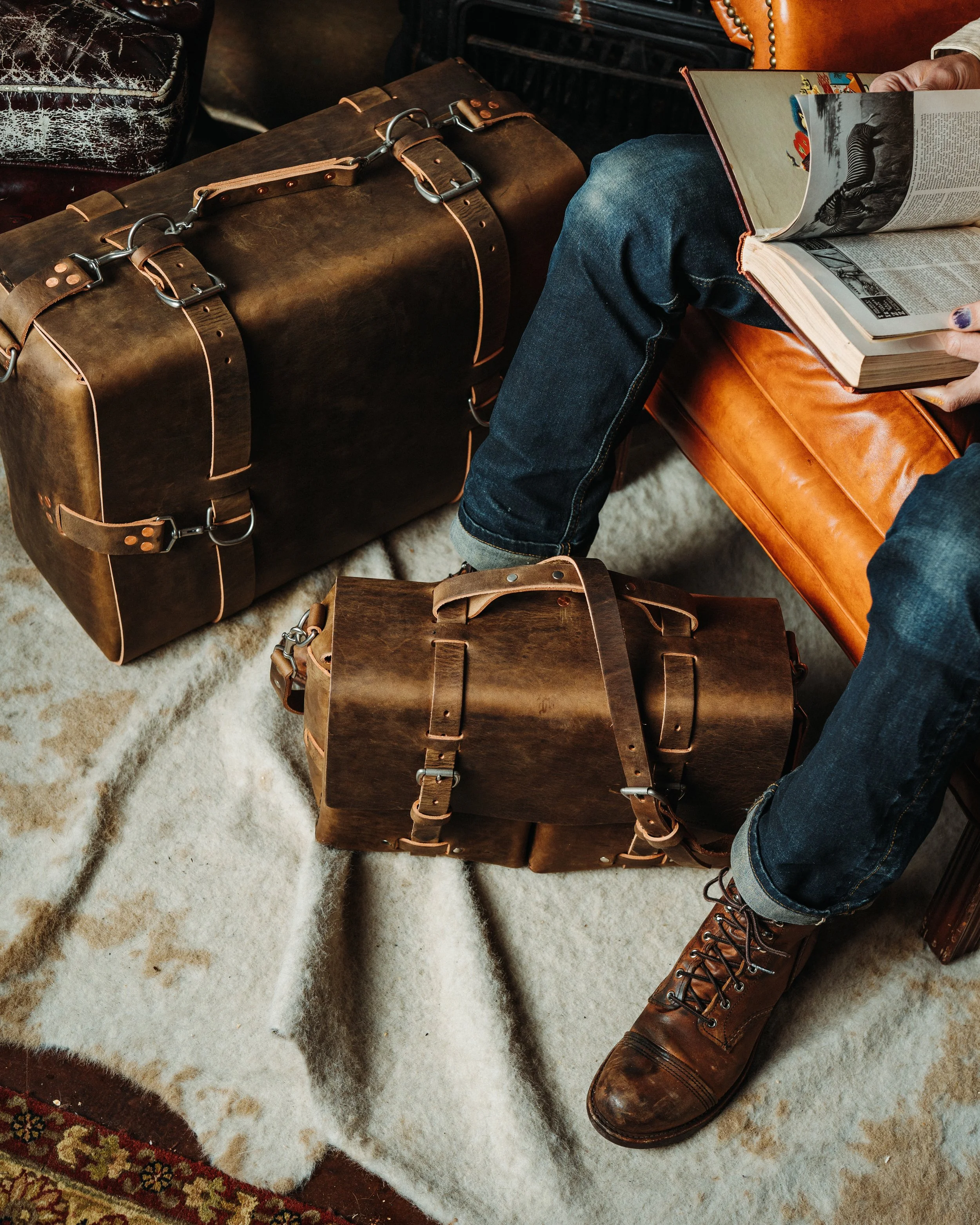 A person wearing brown leather boots and blue jeans sitting on a brown leather couch, reading a magazine. Vintage brown leather suitcases are placed on a fluffy, light-colored rug, with part of a distressed black leather chair visible in the background.