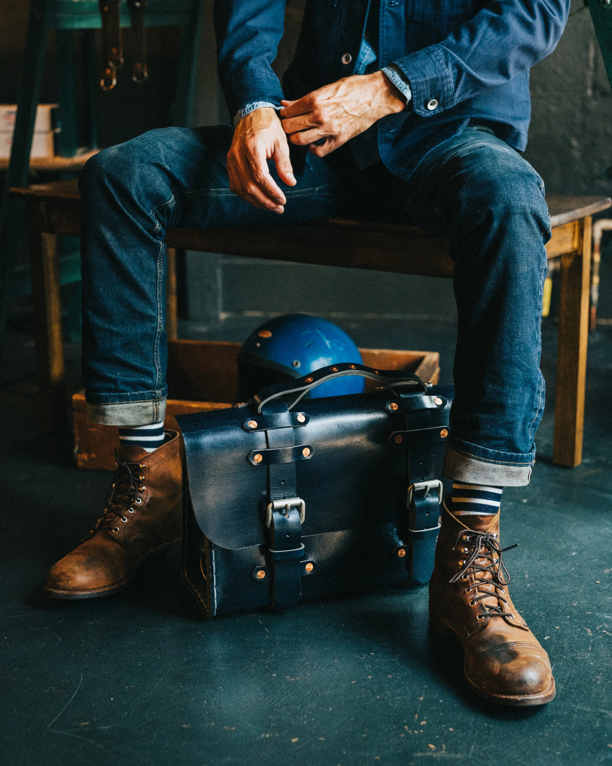 A person wearing brown work boots, dark blue jeans, and a denim jacket is sitting on a wooden bench. They are holding a wheel or helmet for roller skates or rollerblades, with a blue helmet on the floor nearby. The background includes a wooden shelf and a dark wall.