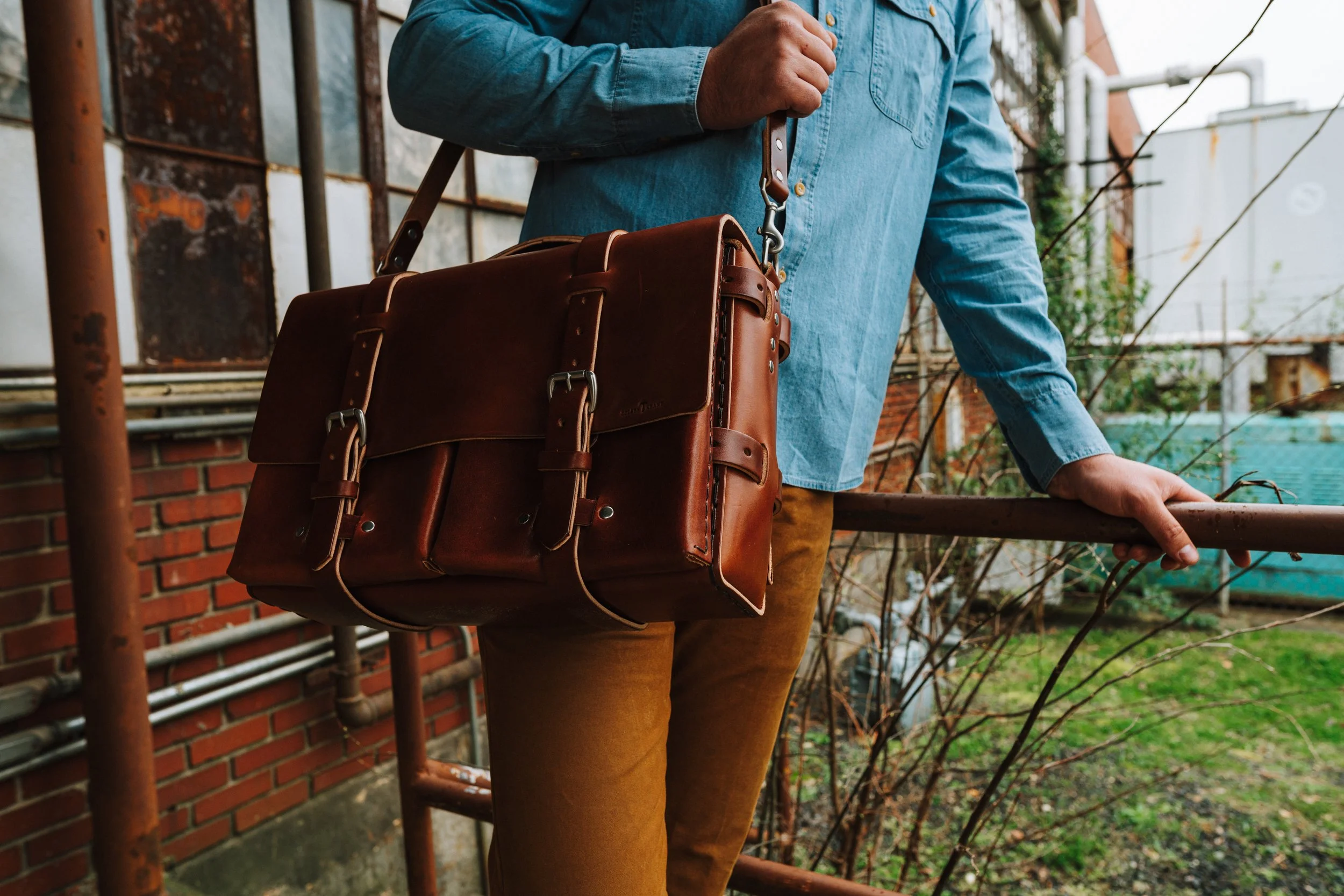 Person in a denim shirt carrying a brown leather bag outdoors near a brick wall and metal pipes.