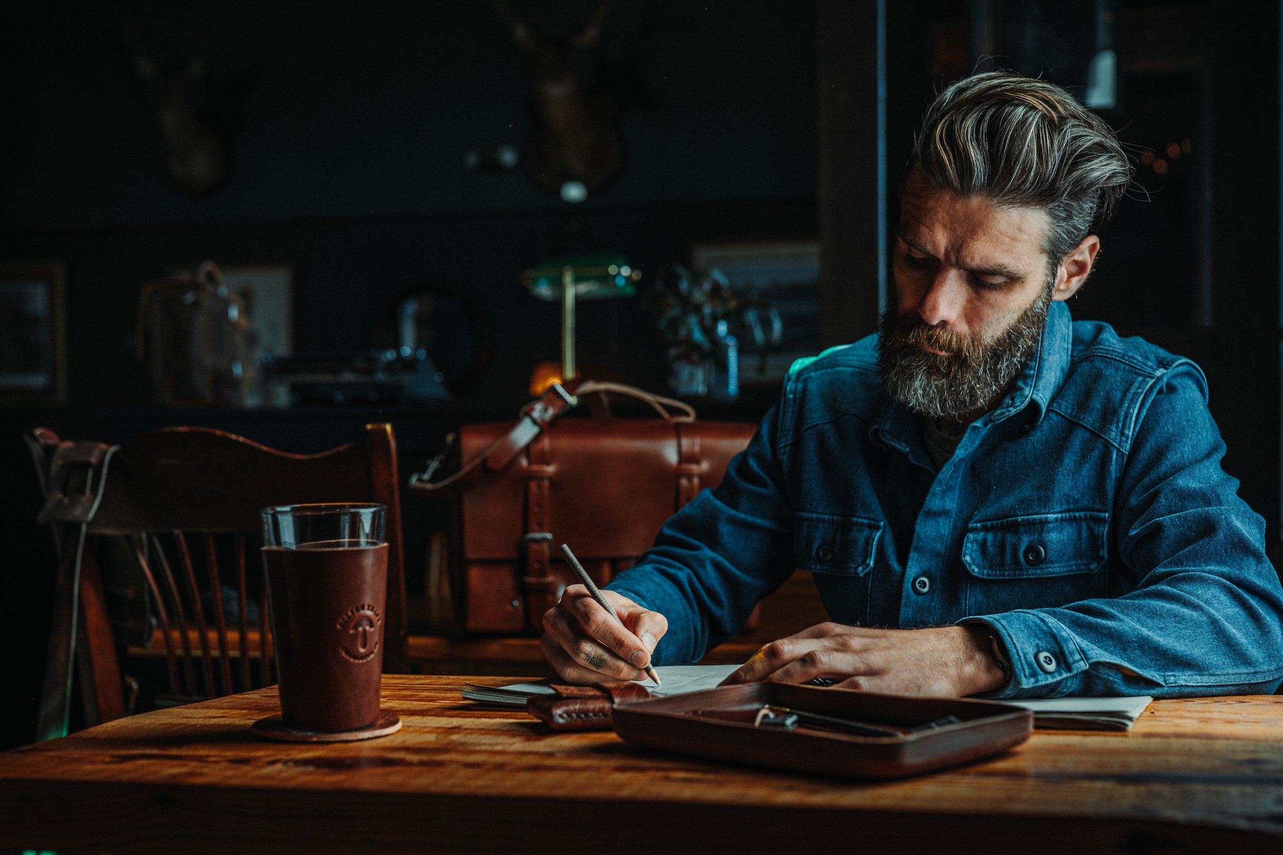 A man with a beard and styled hair, wearing a denim jacket, is sitting at a wooden table writing or drawing on paper in a dimly lit setting.