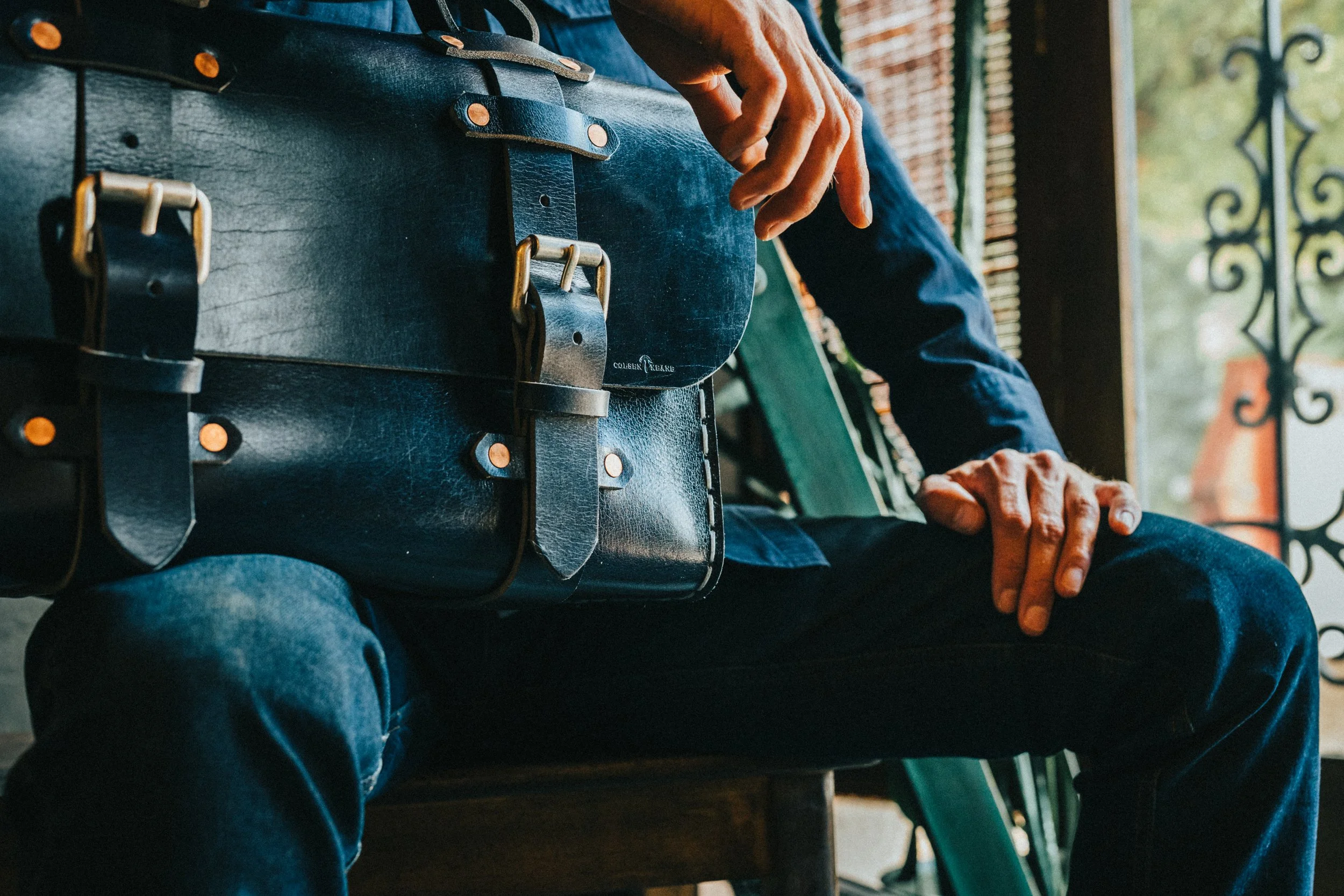 Close-up of a person sitting, with their hand resting on their lap, wearing a dark jacket and blue jeans. The person has a black leather satchel with metal buckles and straps resting on their lap. The background shows a decorative metal railing and a porch or balcony setting.