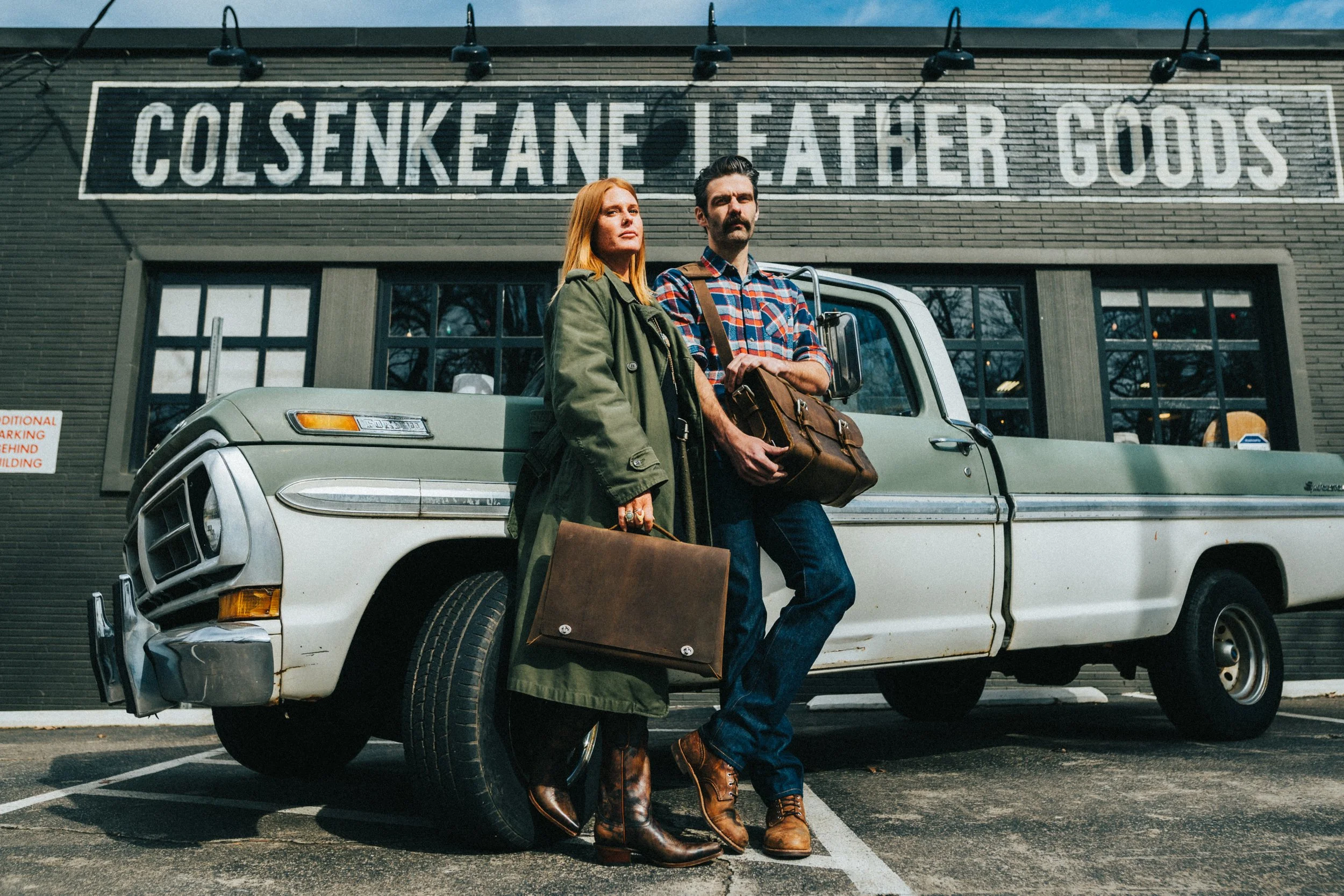 A man and a woman standing in front of a vintage pickup truck outside a brick storefront with a painted sign that reads 'ColsenKeane Leather Goods'.