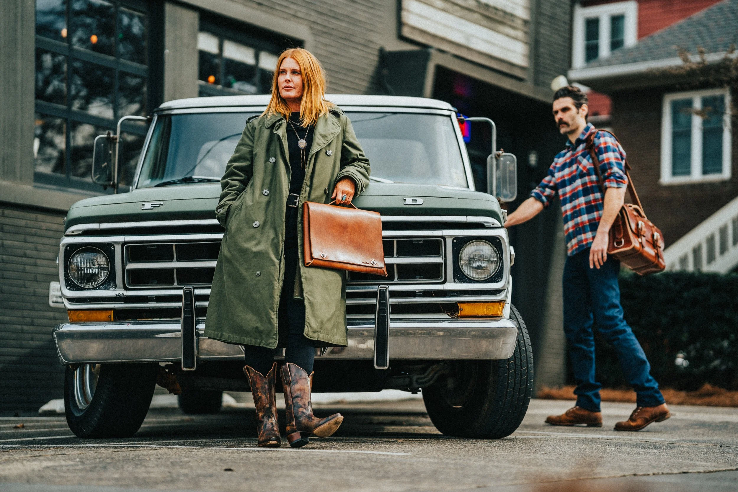 A woman with red hair wearing a green coat and cowboy boots standing in front of a vintage green Ford truck, with a man in a plaid shirt and carrying a leather bag nearby, in an urban residential area.