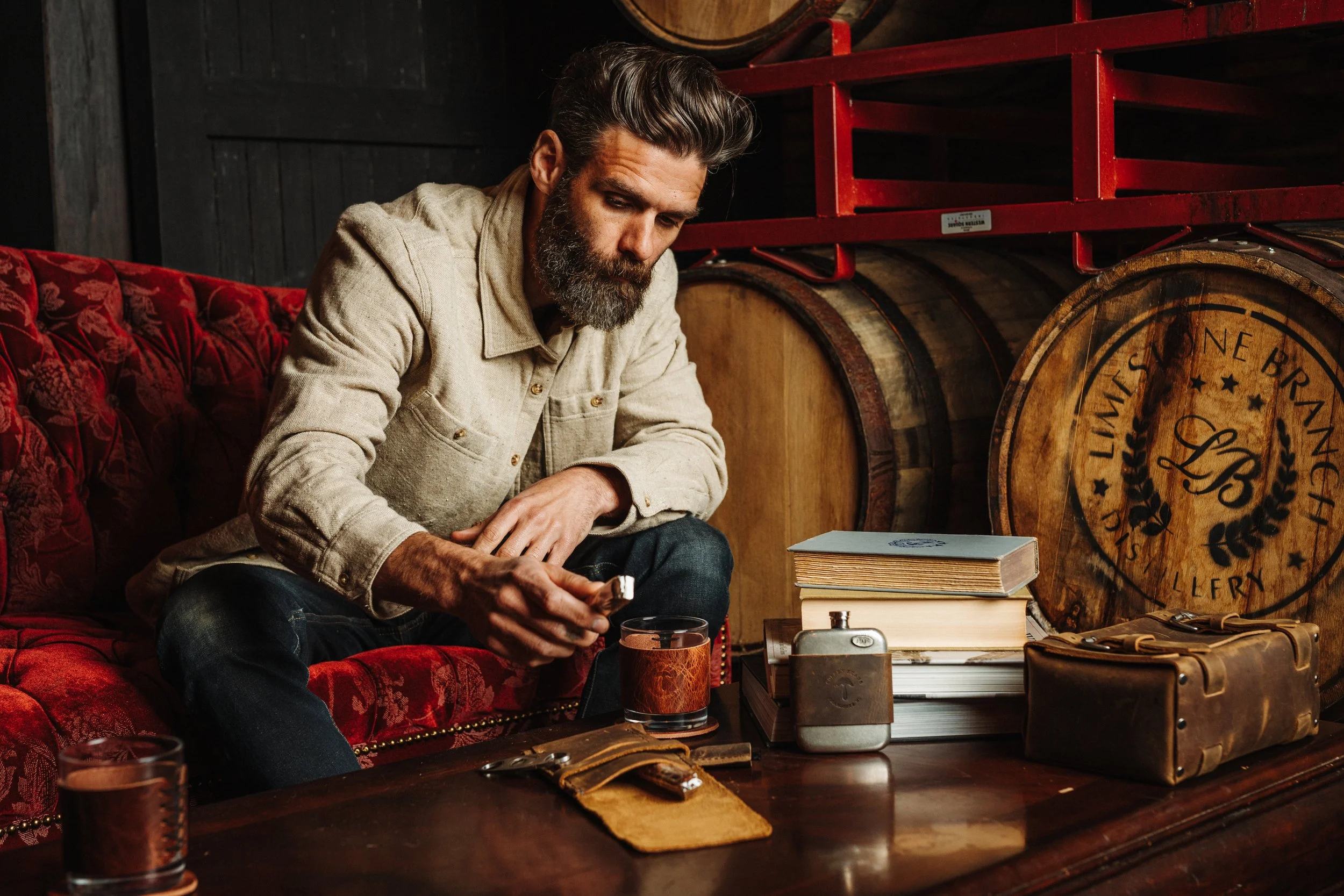 A man with a beard and styled hair sitting on a red cushioned vintage sofa, reading a book in a rustic room with wooden barrels and furniture, with a glass of whiskey on the table.