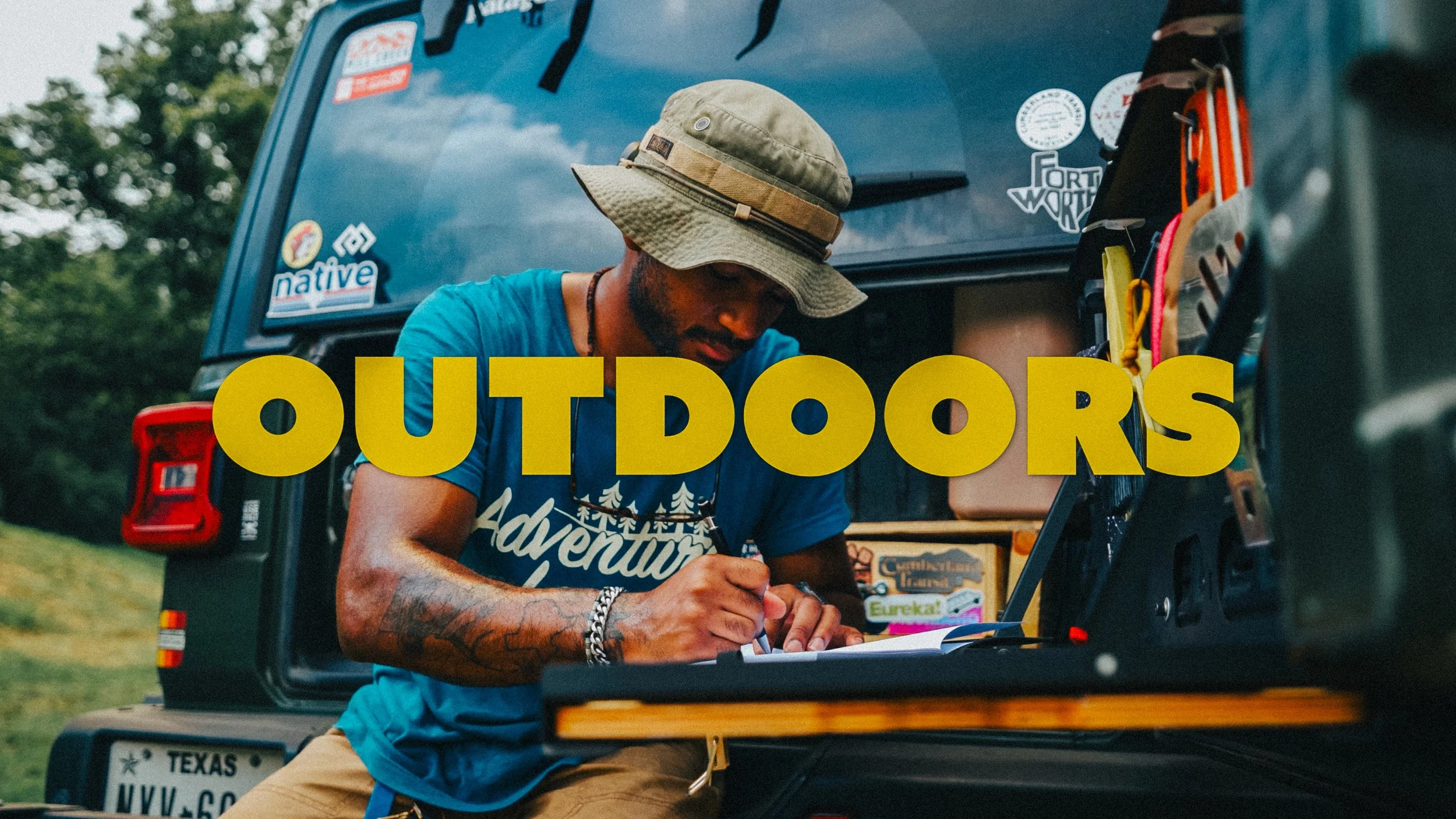 A man wearing a beige bucket hat and a blue t-shirt with white text is sitting in the open trunk of a vehicle, writing or drawing on a paper. The vehicle is decorated with various stickers, and the background shows a grassy outdoor area with trees. The word 'OUTDOORS' is overlaid in large yellow letters across the bottom of the image.