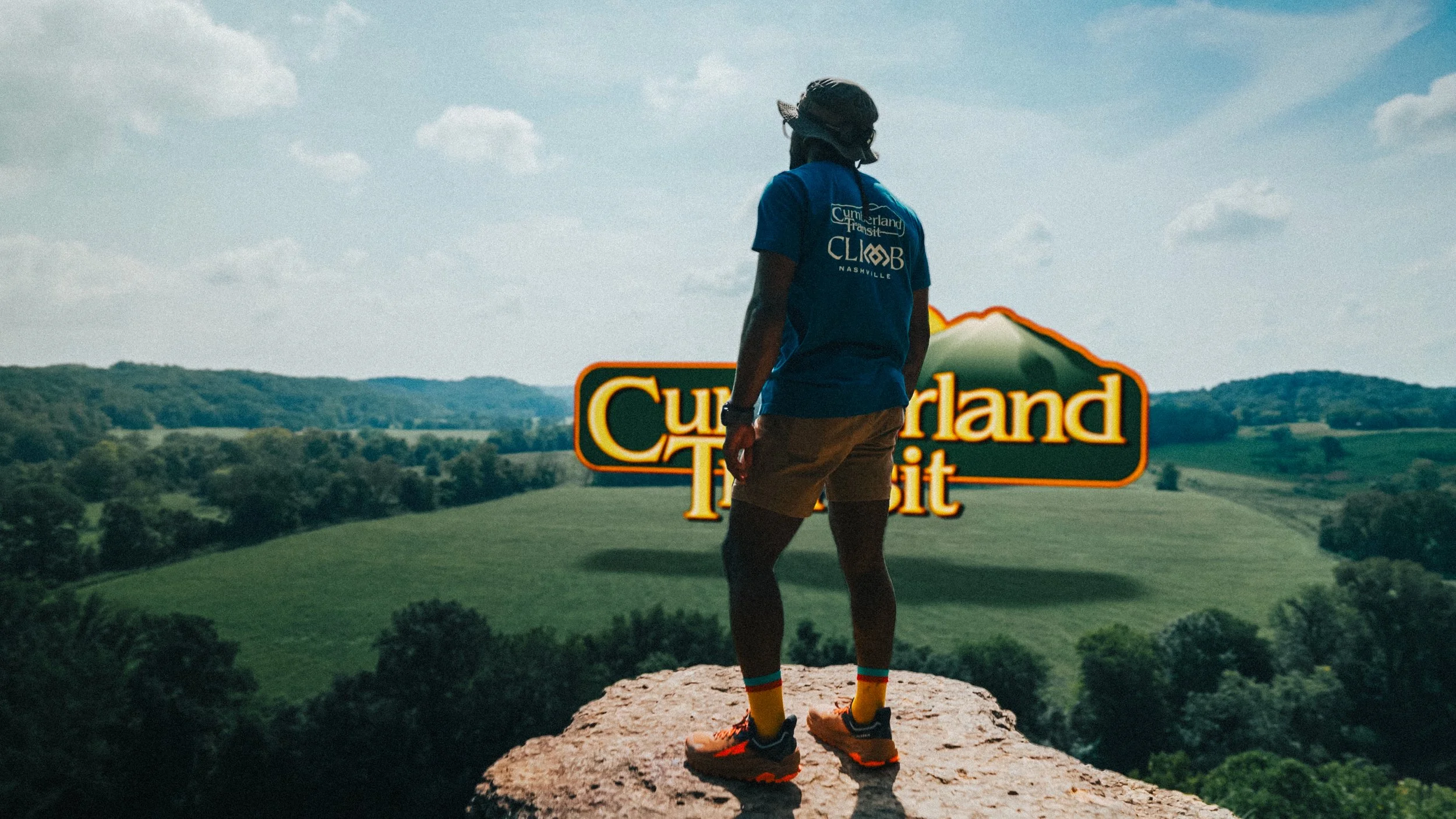 Person standing on a rock overlooking a landscape with hills and trees, with a large Cumberland Transit sign in the distance.