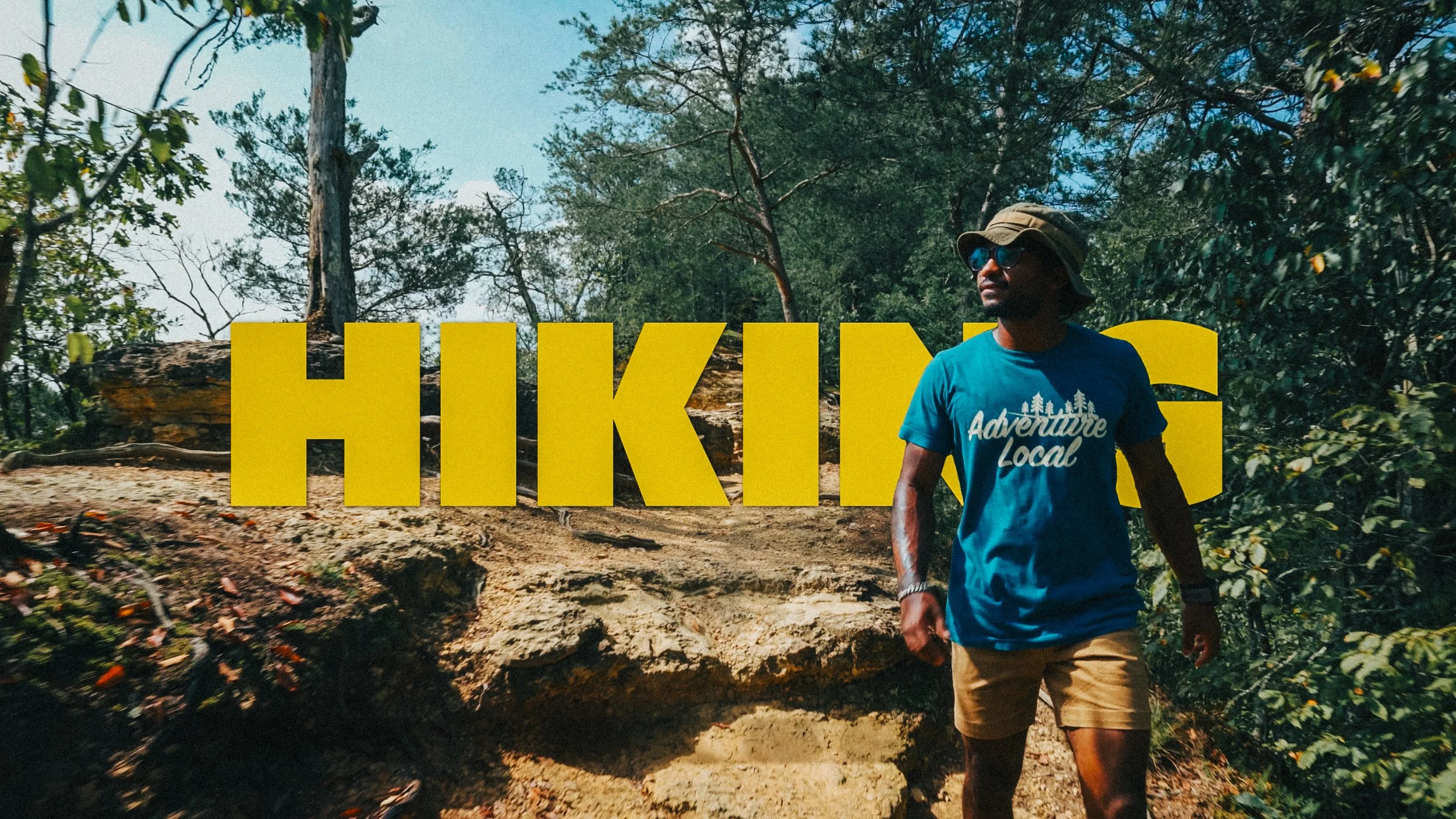 A man wearing a blue t-shirt with 'Adventure Local' printed on it, a wide-brimmed hat, sunglasses, and beige shorts, walking past a large yellow sign that says 'HIKING' in a forested area.