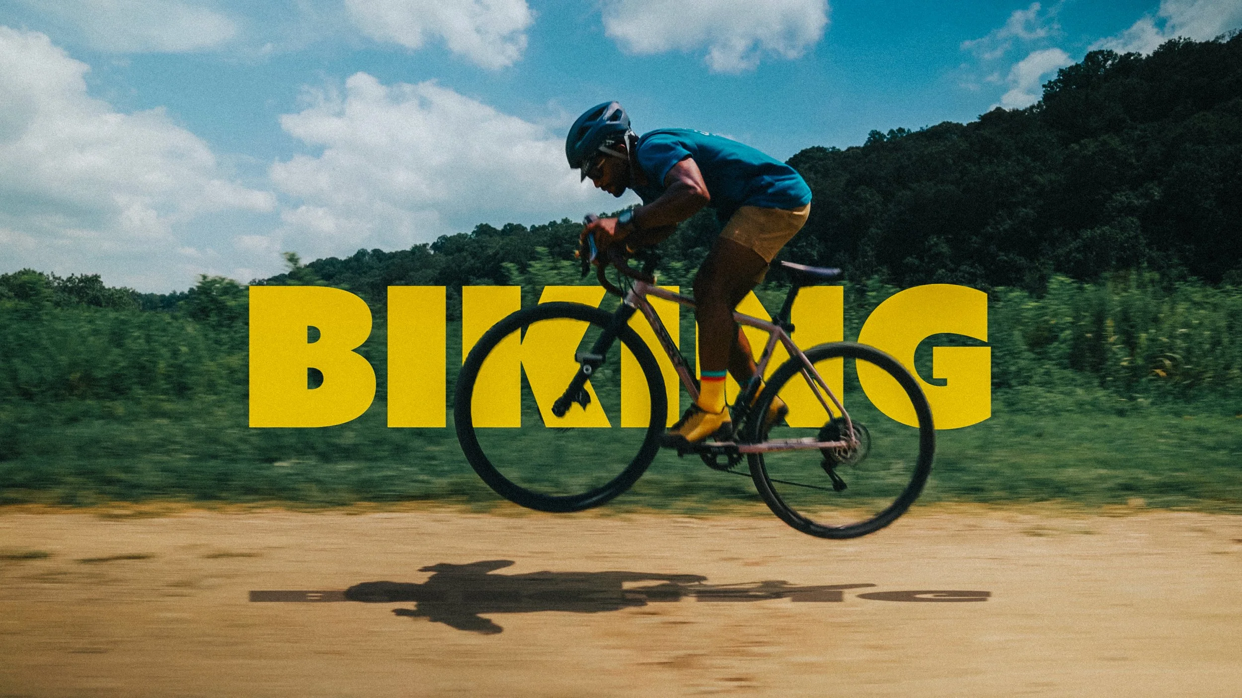 A person wearing a blue helmet and shirt riding a bike on a dirt trail with green trees and hills in the background, with the word 'BIKING' in large yellow letters overlaying the image.