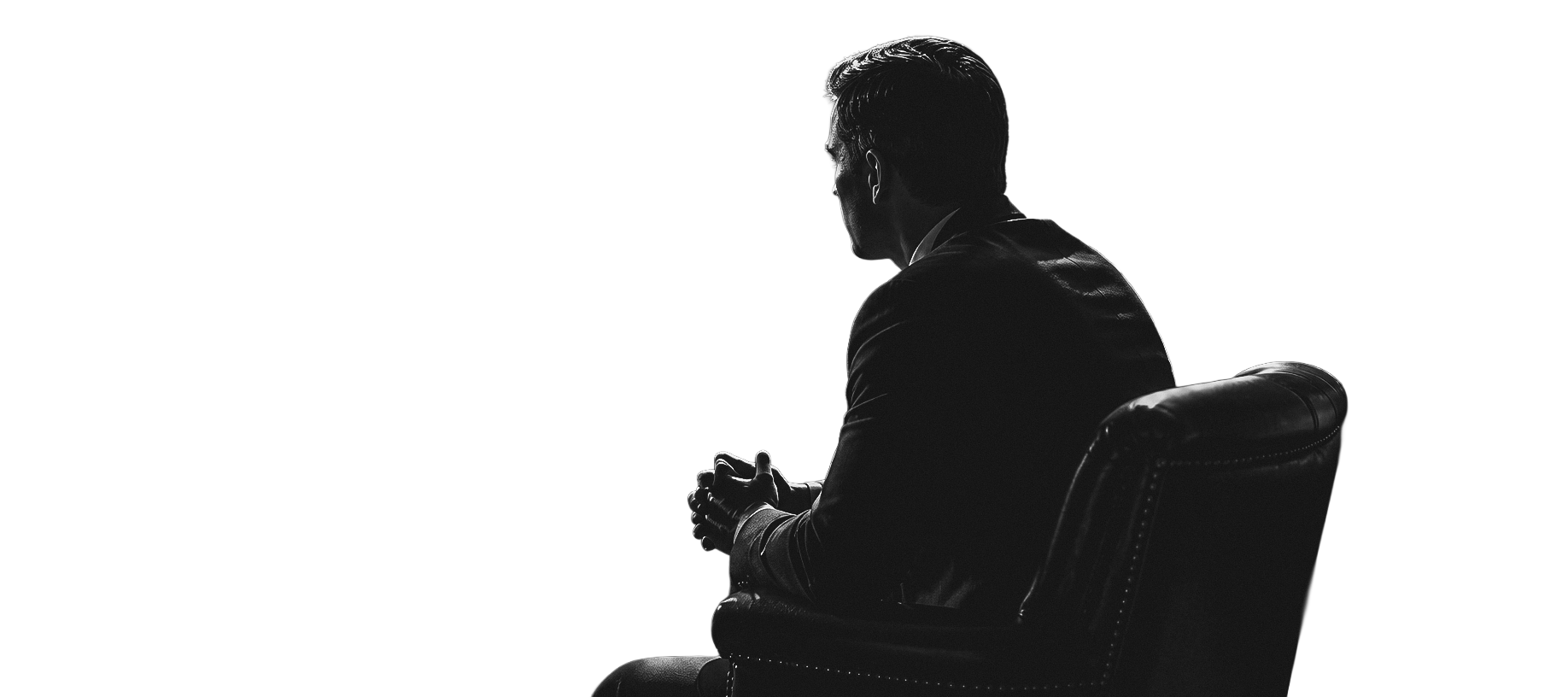 Black and white photo of a man sitting in a leather chair, wearing a suit, with a dark background and a gradient light source behind him.