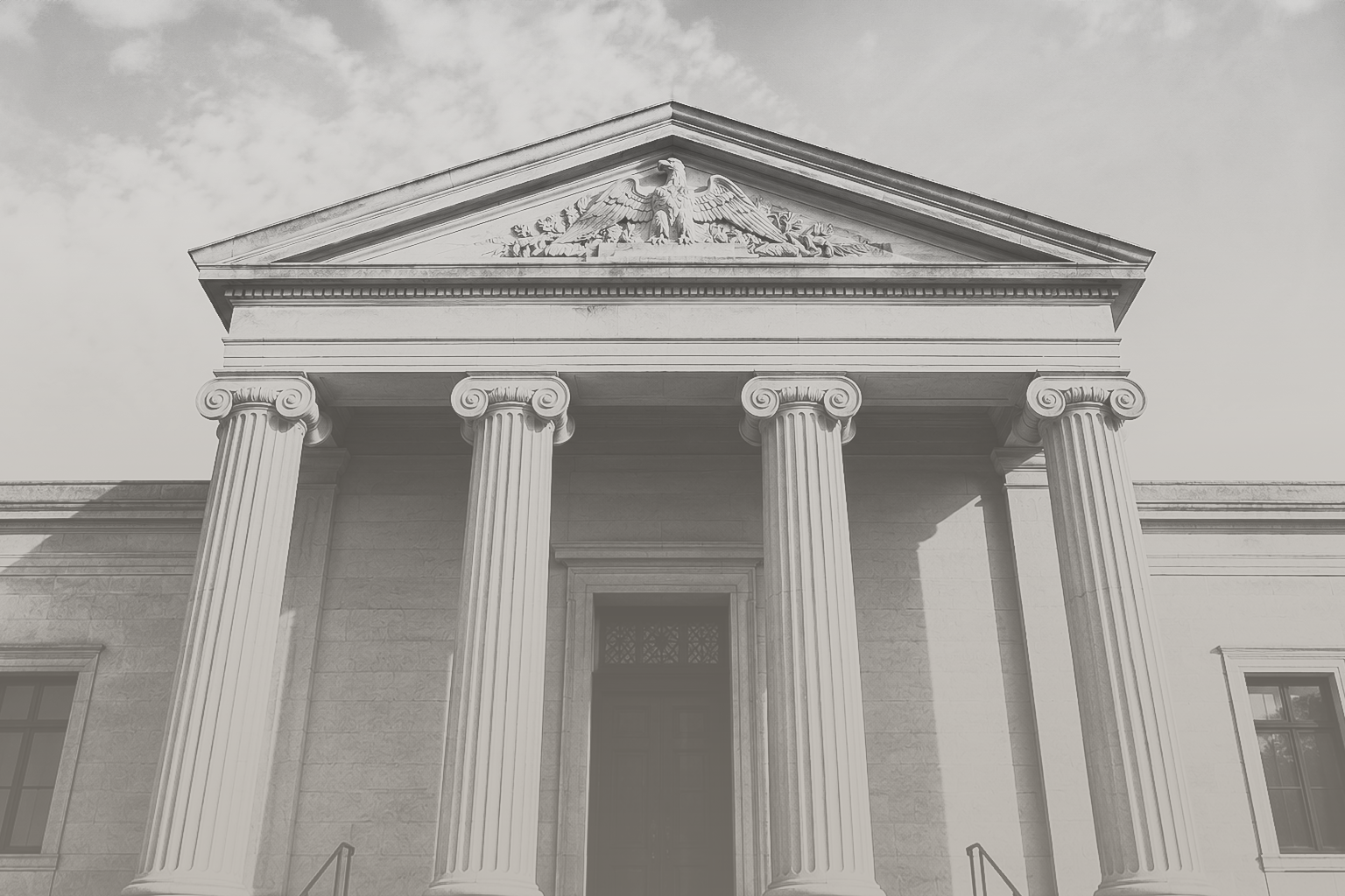 A grayscale photograph of a neoclassical building front with four large columns supporting a triangular pediment with an eagle sculpture at the center.